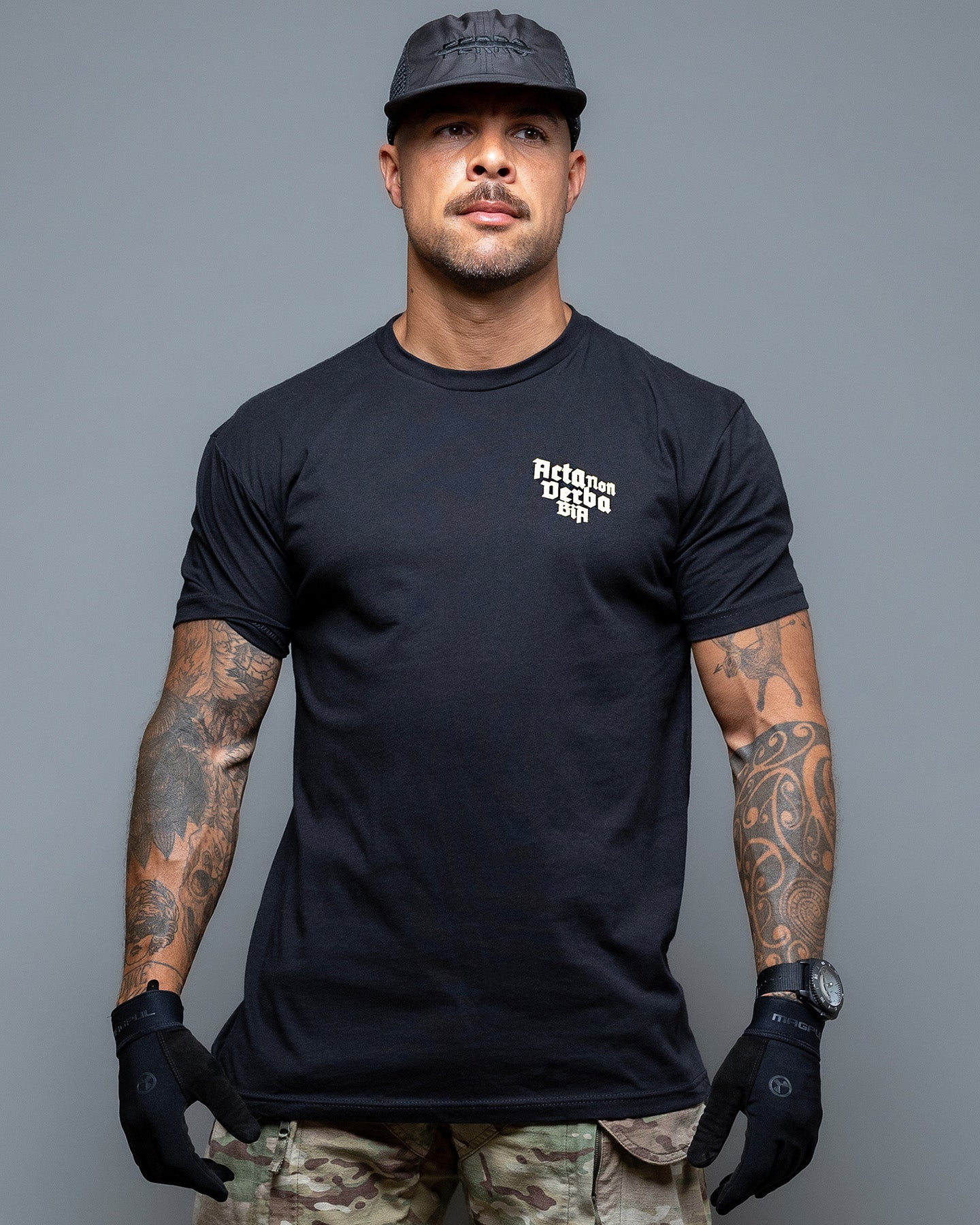 Ein Mann mit schwarzer Kappe und sichtbaren Tattoos unter seinem Acta Non Verba T-Shirt von Brothers in Arms steht selbstbewusst vor einem grauen Hintergrund. Das Funktionsshirt und die Handschuhe betonen „Acta Non Verba“, das in jede Faser seiner atmungsaktiven Kleidung eingraviert ist. #color_black