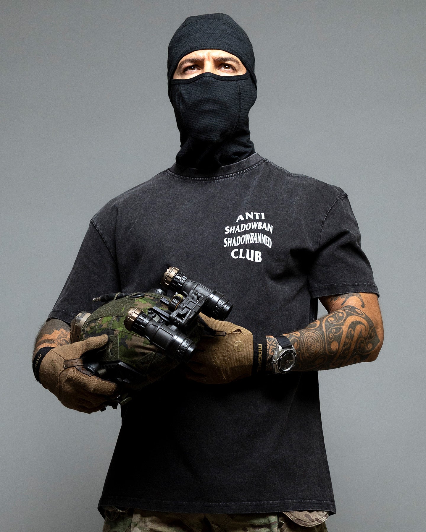 Person wearing a black balaclava and gloves, holding tactical gear, with a black t-shirt featuring text. #color_black