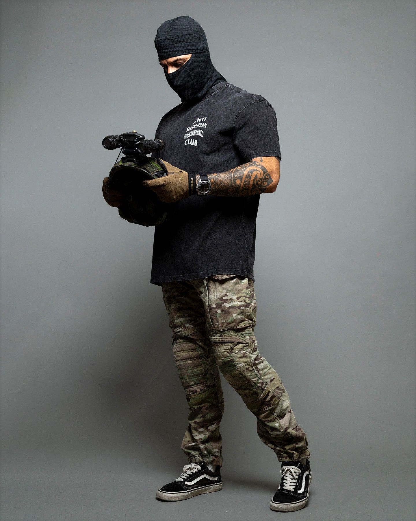 Person wearing a black balaclava, black t-shirt, camouflage pants, and black sneakers on a gray background. #color_black