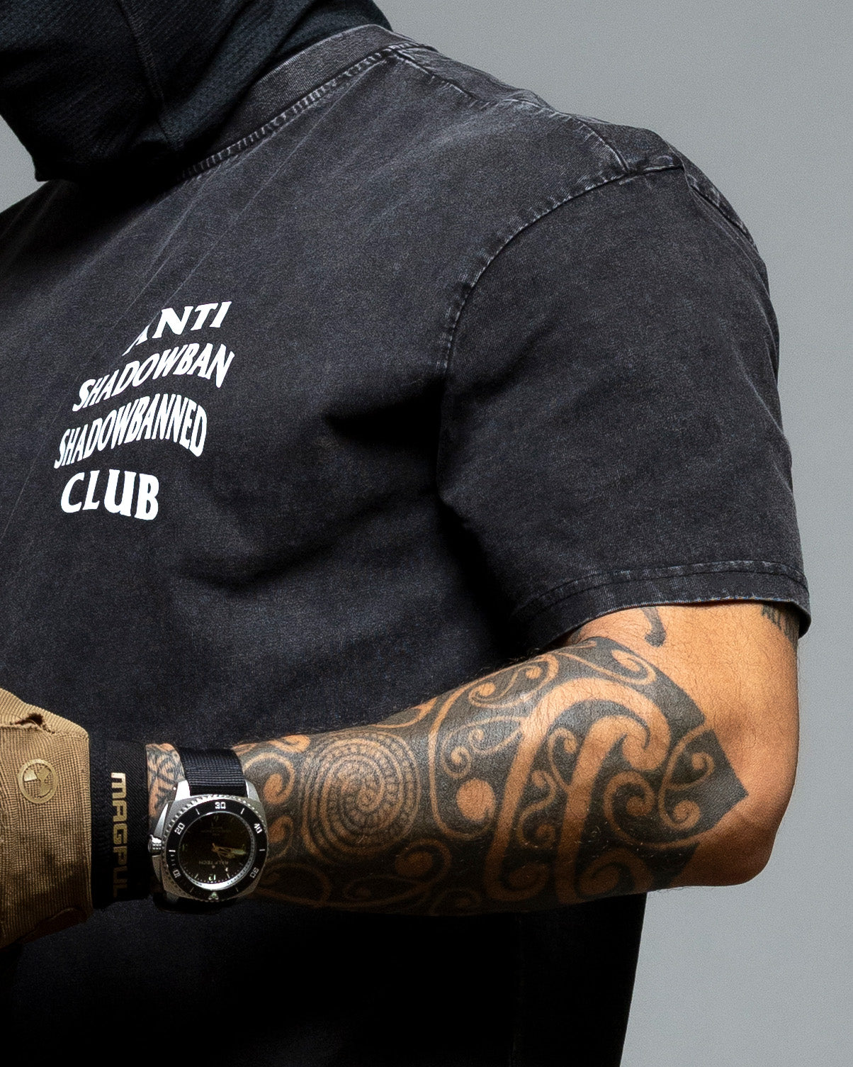 Person wearing a black shirt with text and showing tattoos on a gray background. #color_black