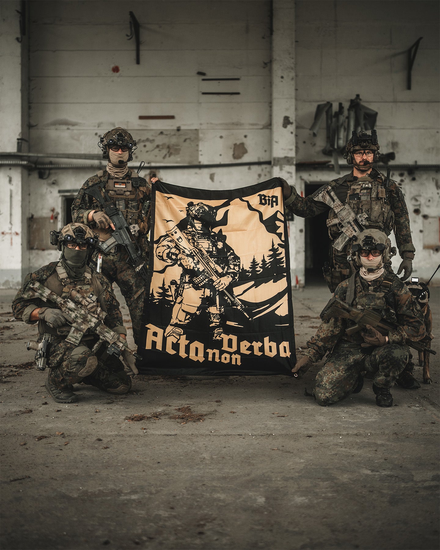 Four individuals in tactical gear holding a banner with 'Acta Non Verba' branding in an industrial setting. #in_field