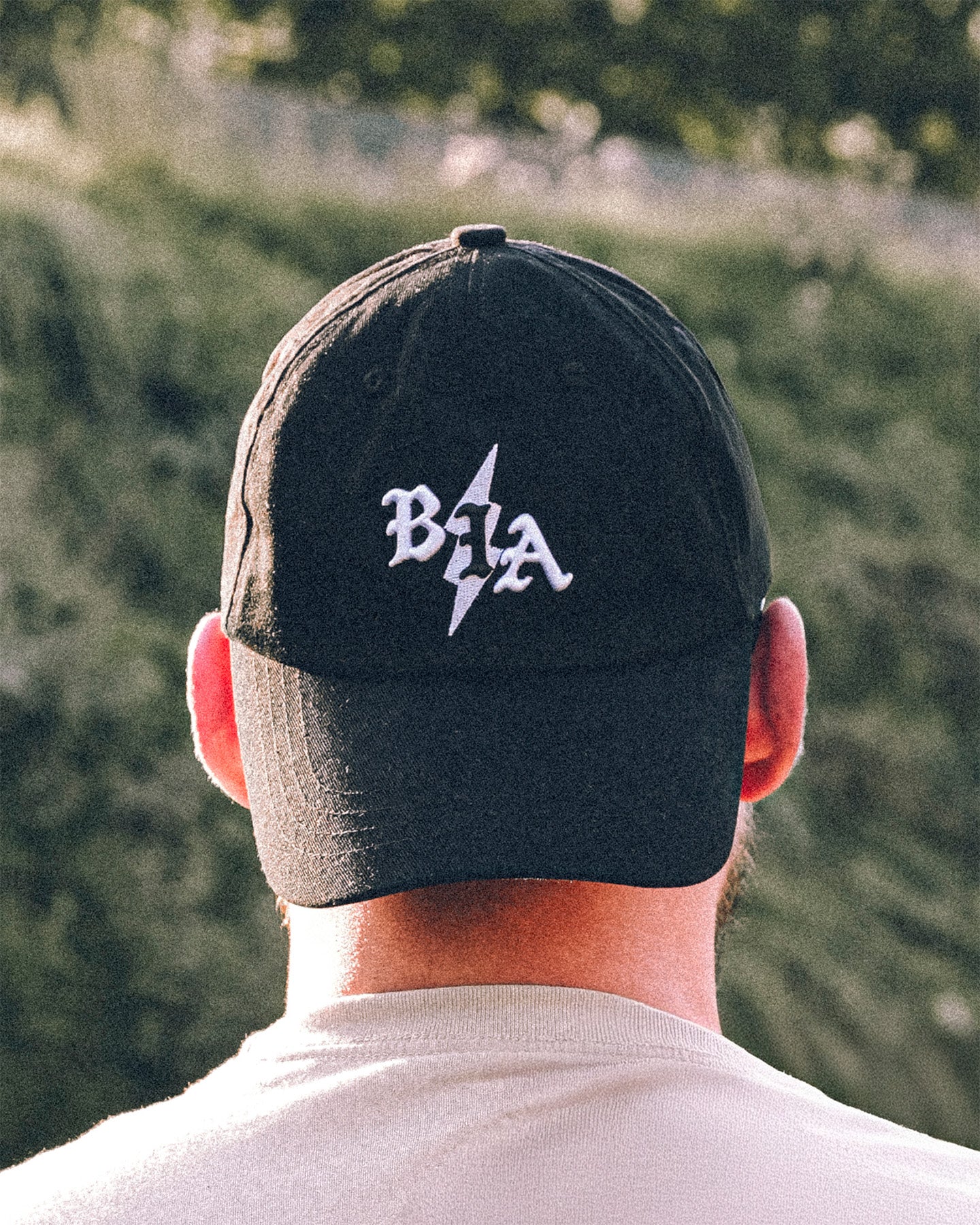 Person wearing a black cap with a logo, facing away from the camera with a blurred green background.