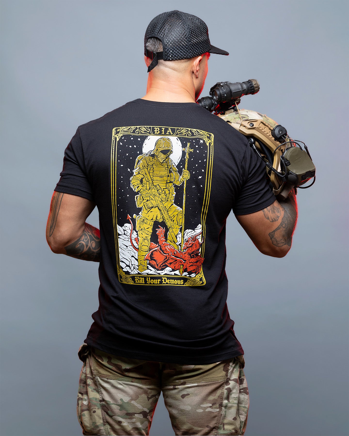 Person wearing a black t-shirt with a graphic design on the back, holding a rifle against a gray background. #color_black