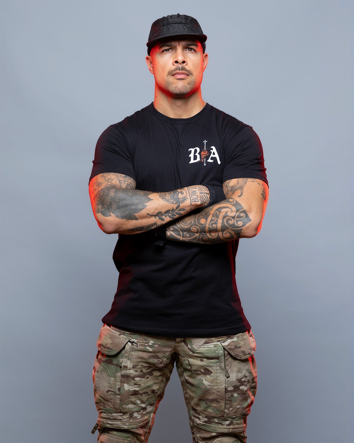 Man wearing a black t-shirt with a logo and camouflage pants against a gray background. #color_black