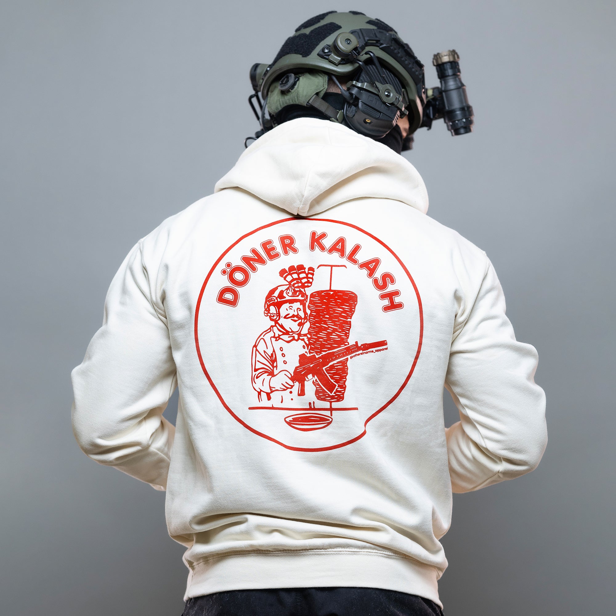 Eine Person trägt einen taktischen Helm und den „Döner Kalash Spicy Hoodie“ von Brothers in Arms mit einer farbenfrohen Döner-Illustration auf der Rückseite. #color_cream