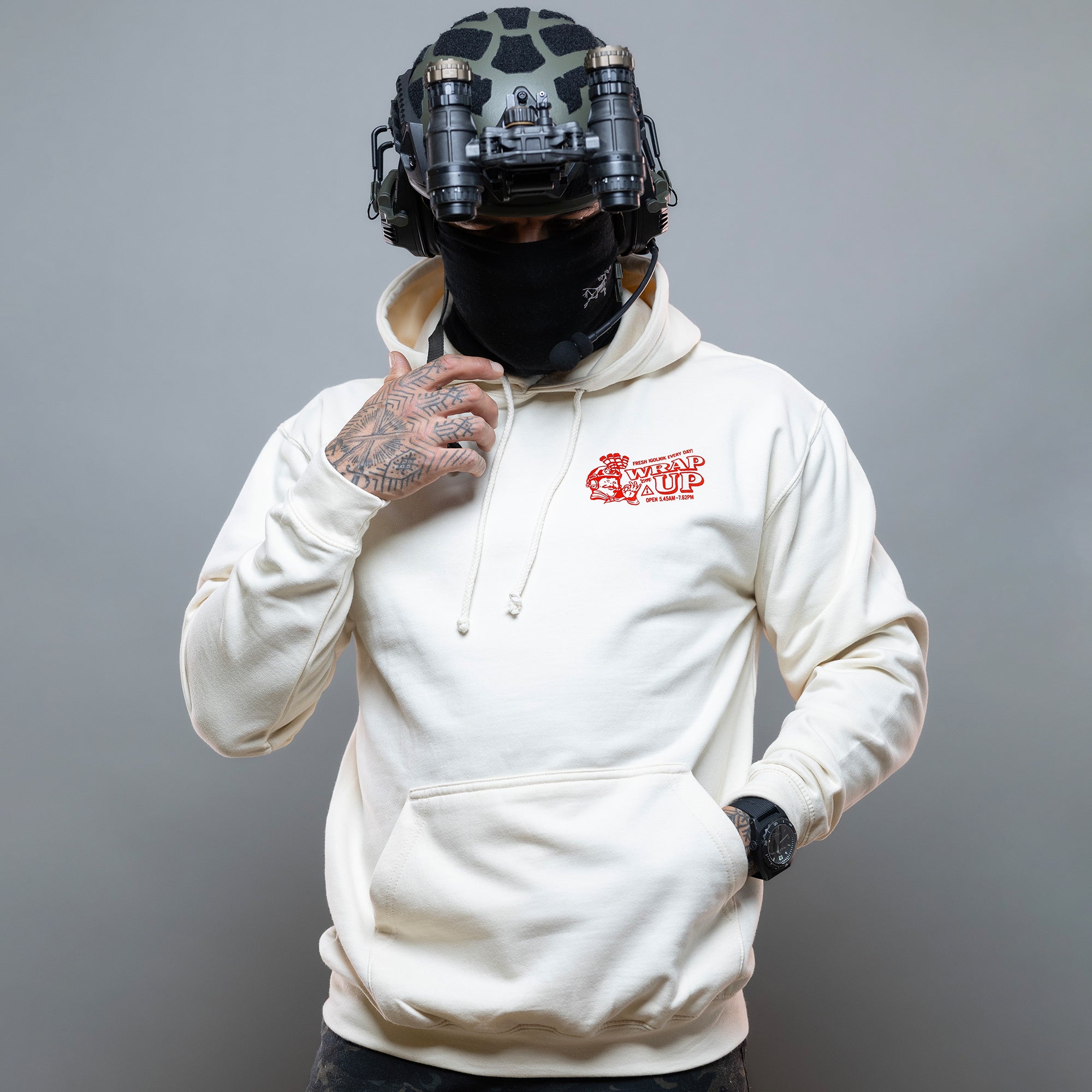 Eine Person steht vor einem grauen Hintergrund und trägt einen Döner Kalash Spicy Hoodie von Brothers in Arms, dazu einen taktischen Helm und eine schwarze Maske. Der Hoodie hat ein auffälliges rotes Logo auf der Brust. #color_cream