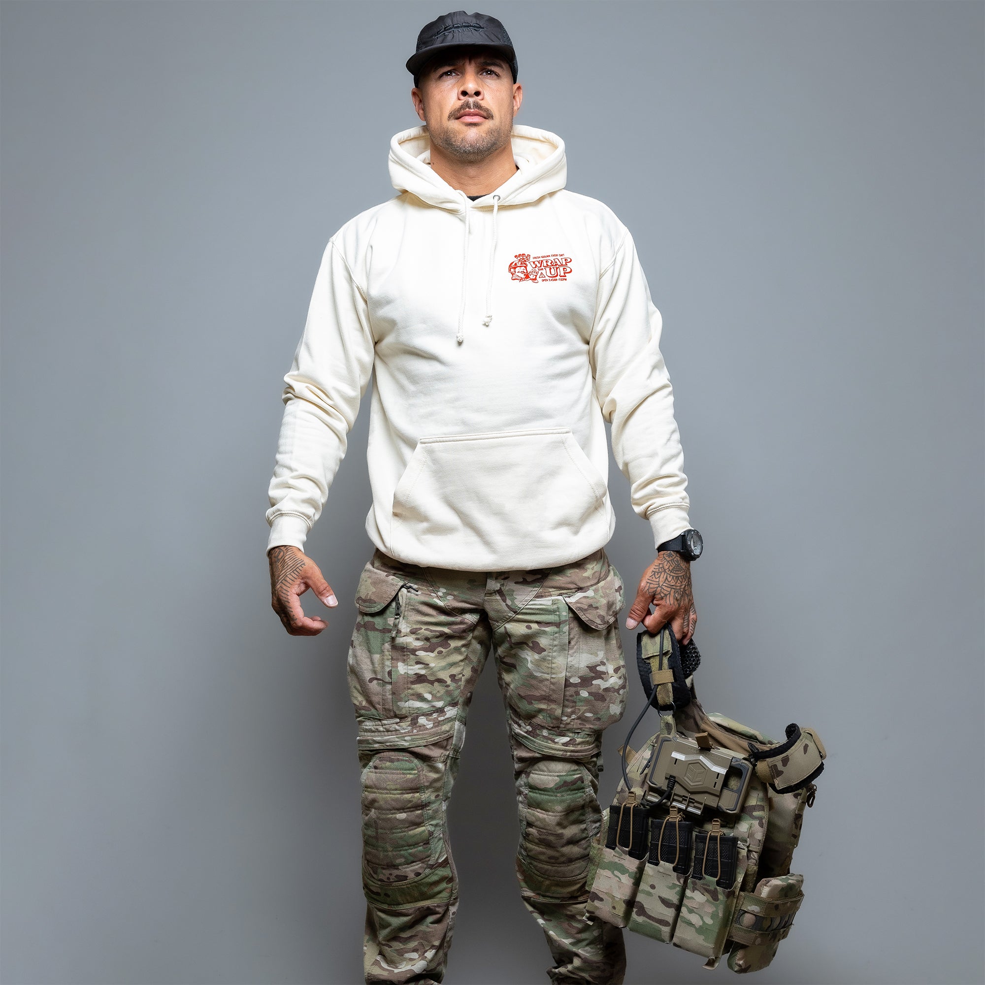 Ein Mann mit einem Döner Kalash Spicy Hoodie von Brothers in Arms und Tarnhosen hält eine taktische Weste und eine schwarze Mütze in der Hand und strahlt vor einem grauen Hintergrund Selbstvertrauen aus, als wäre er bereit, die Streetstyle-Szene zu erobern. #color_cream