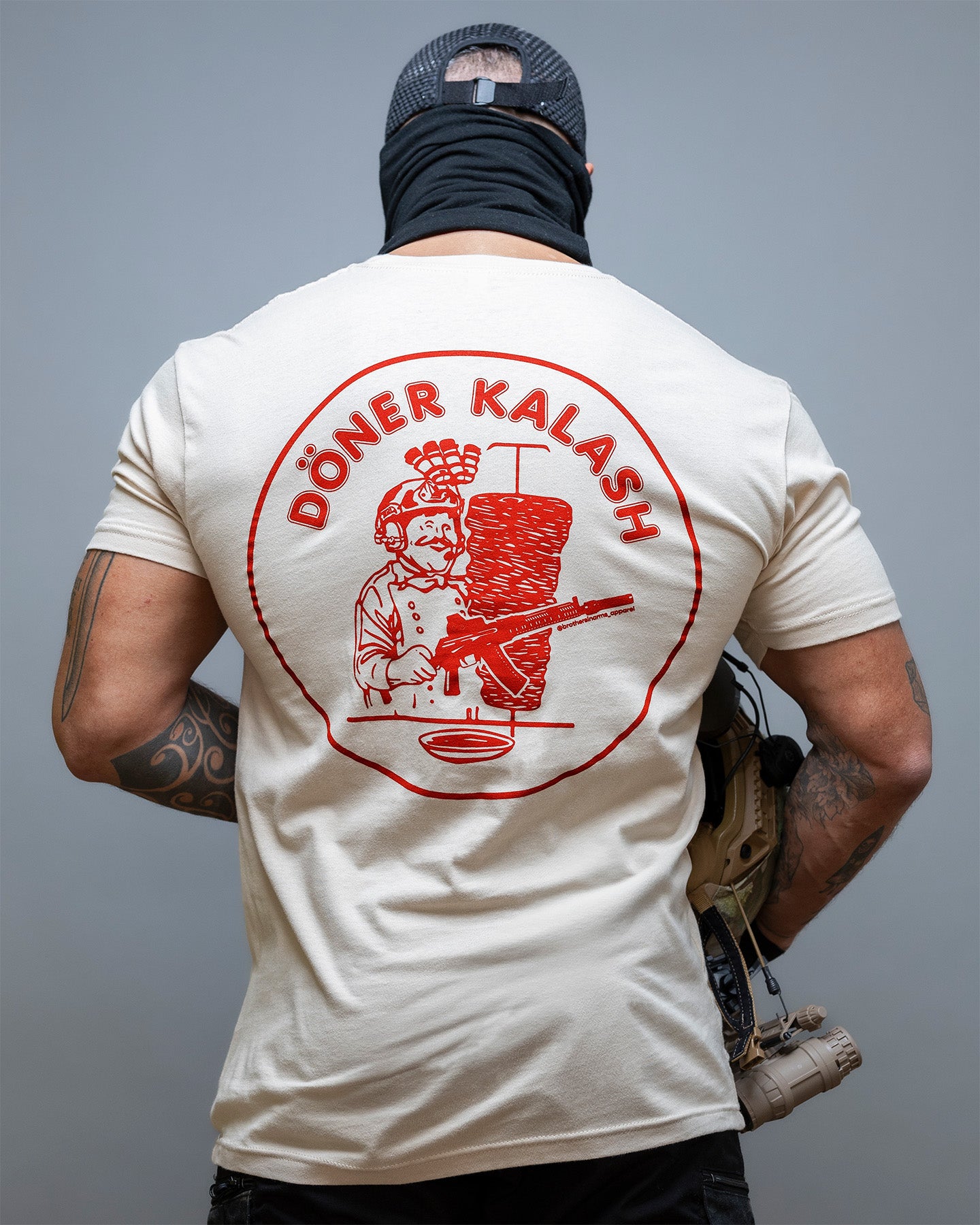 Person wearing a tan t-shirt with a red graphic and text on the back, standing against a gray background. #color_sand