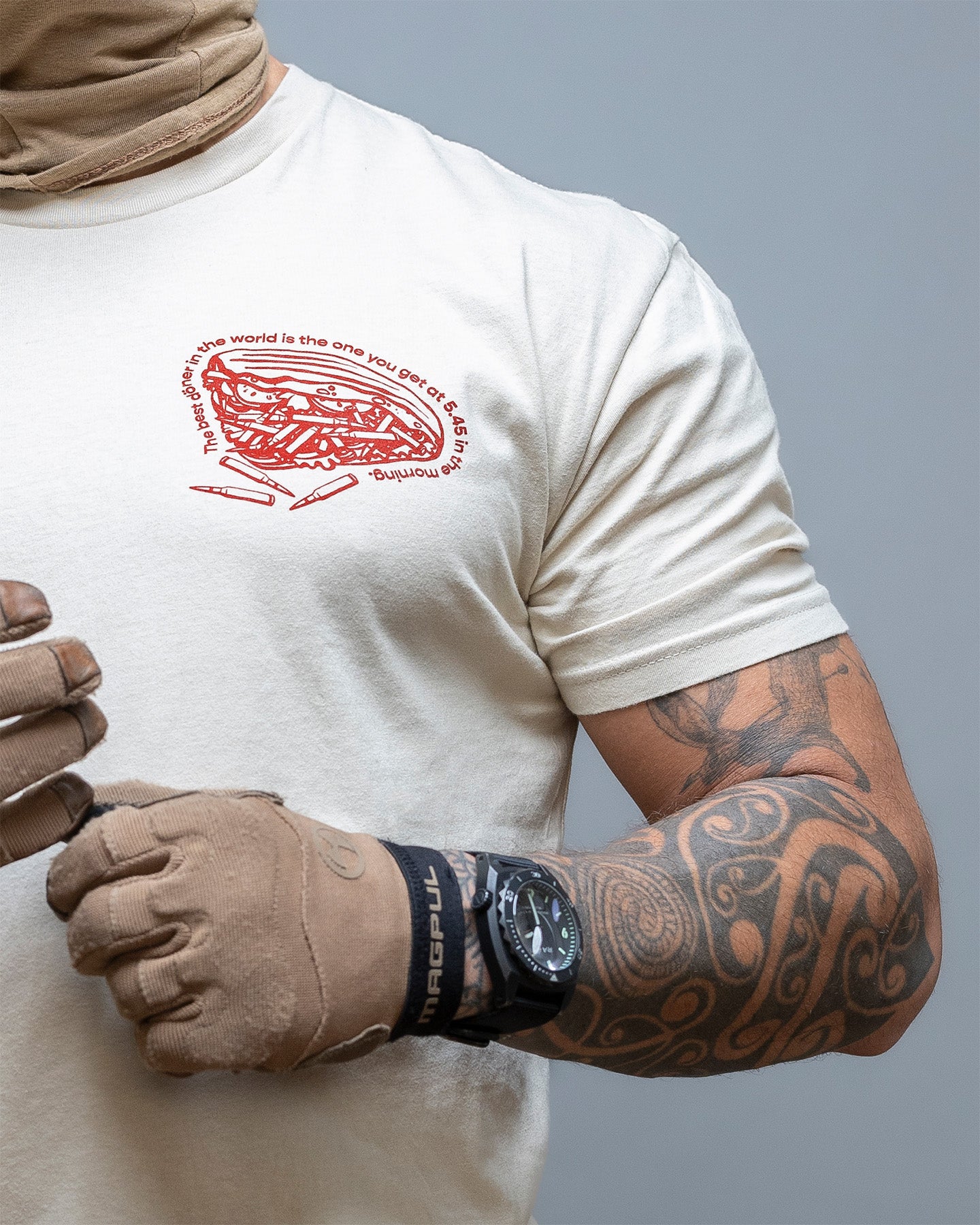 Person wearing a white t-shirt with a red graphic design, brown gloves, and a tattooed arm. #color_sand