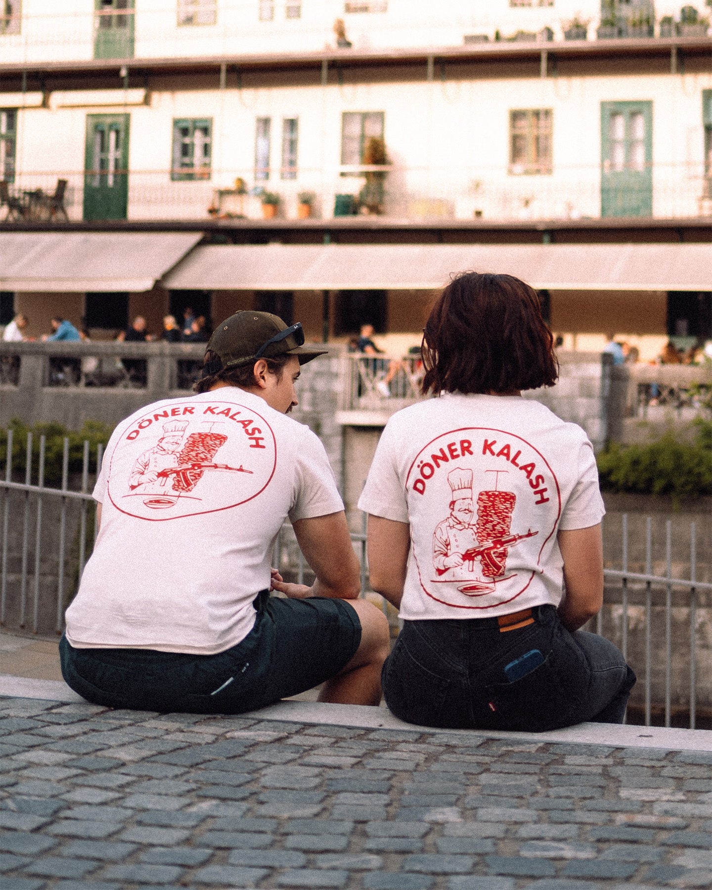 Two people sitting on a ledge wearing 'Döner Kalash' t-shirts. #color_white #in_field