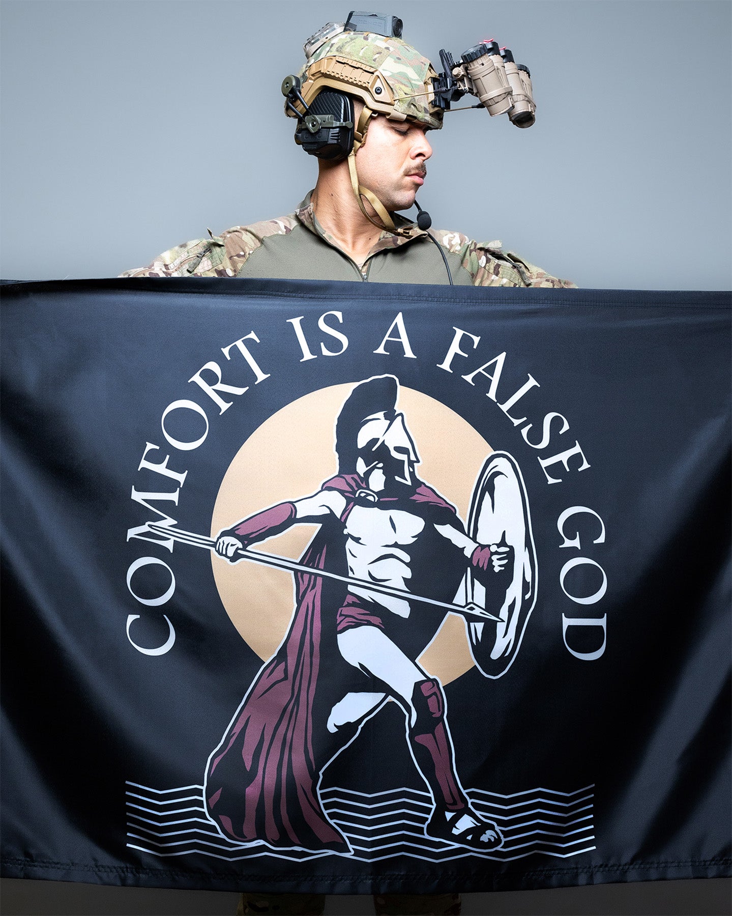A person in military camouflage attire holding up a black flag with a Spartan warrior illustration and the text 'COMFORT IS A FALSE GOD'.