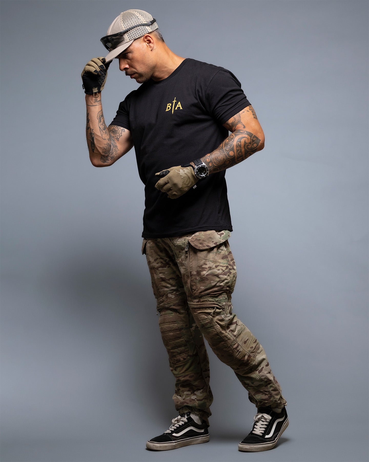 Man wearing a black t-shirt with a logo, camouflage pants, and black sneakers on a gray background. #color_black