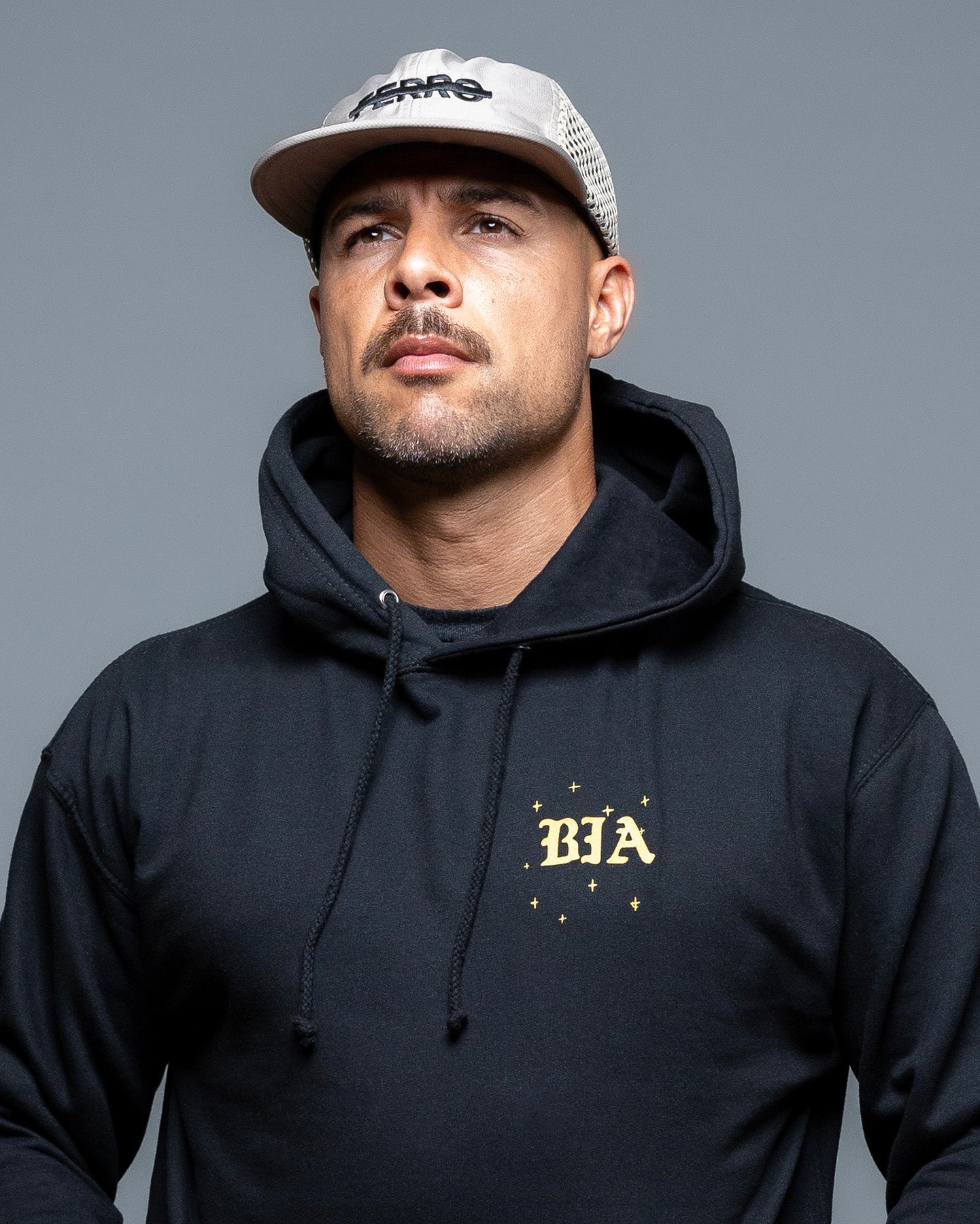 Man wearing a black hoodie with 'BIA' logo and a cap on a gray background. #color_black