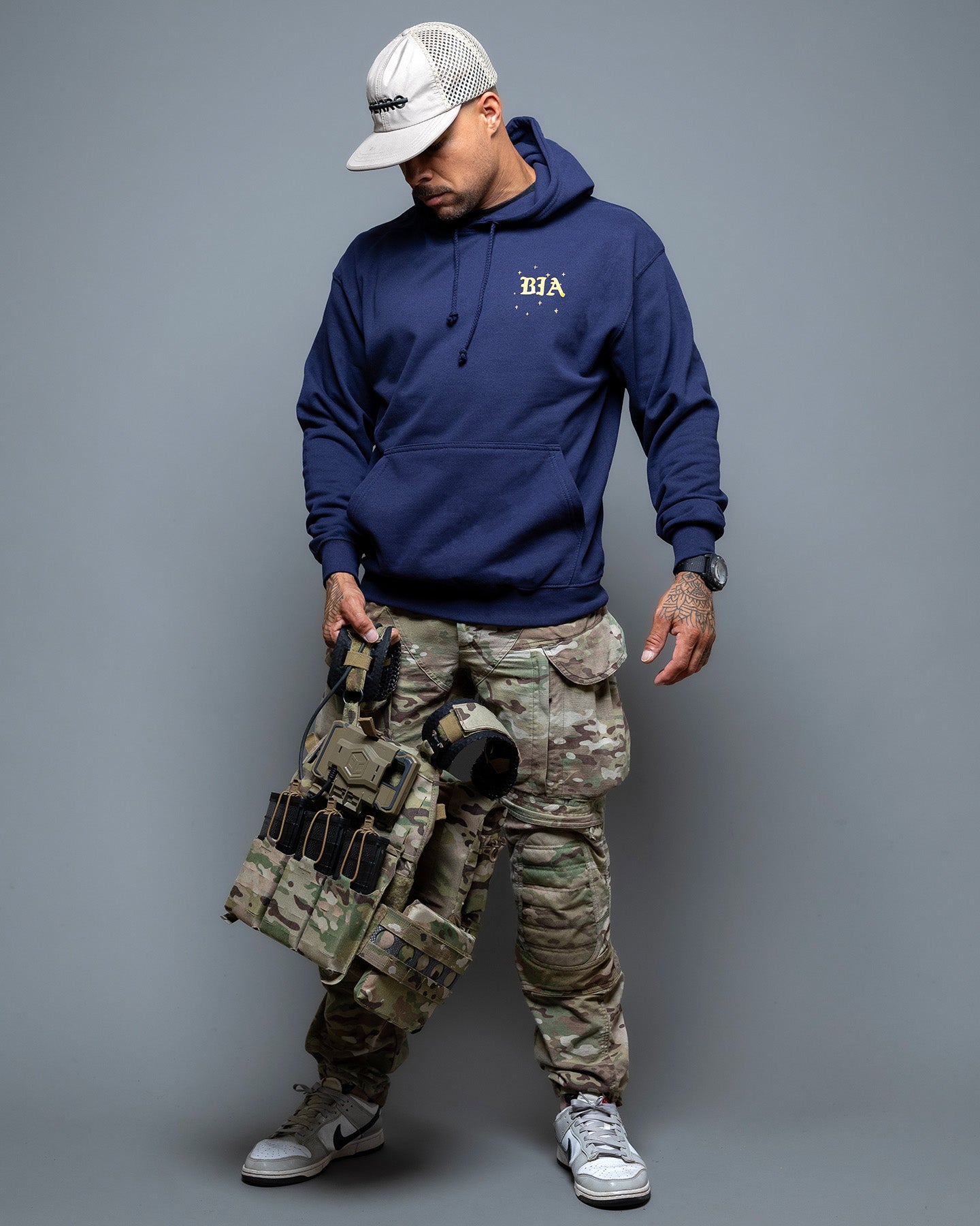 Person wearing a navy hoodie with a logo, camouflage pants, and tactical gear on a gray background. #color_navy