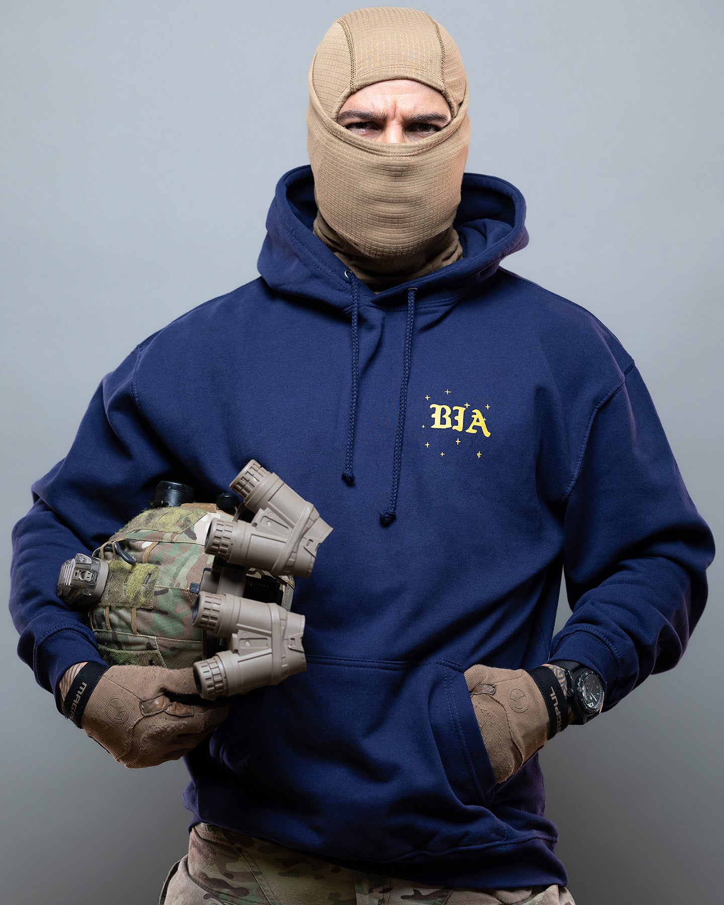 Person wearing a blue hoodie with 'BIA' logo, holding tactical gear against a gray background. #color_navy