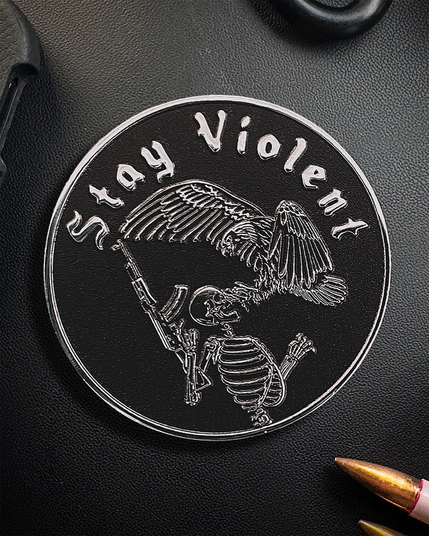 Circular Coin with skeleton and eagle design and 'Stay Violent' text on a black surface.