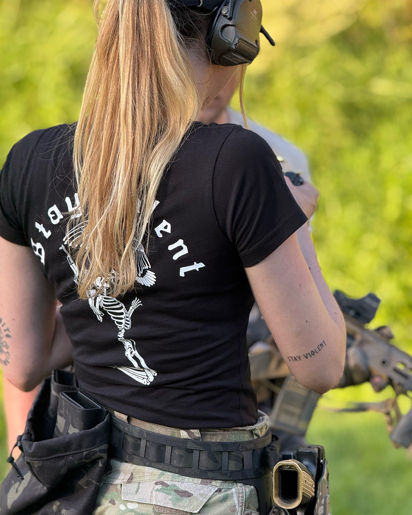 Person wearing a black t-shirt with text and graphics, holding a rifle in a grassy outdoor setting. #color_black #in_field