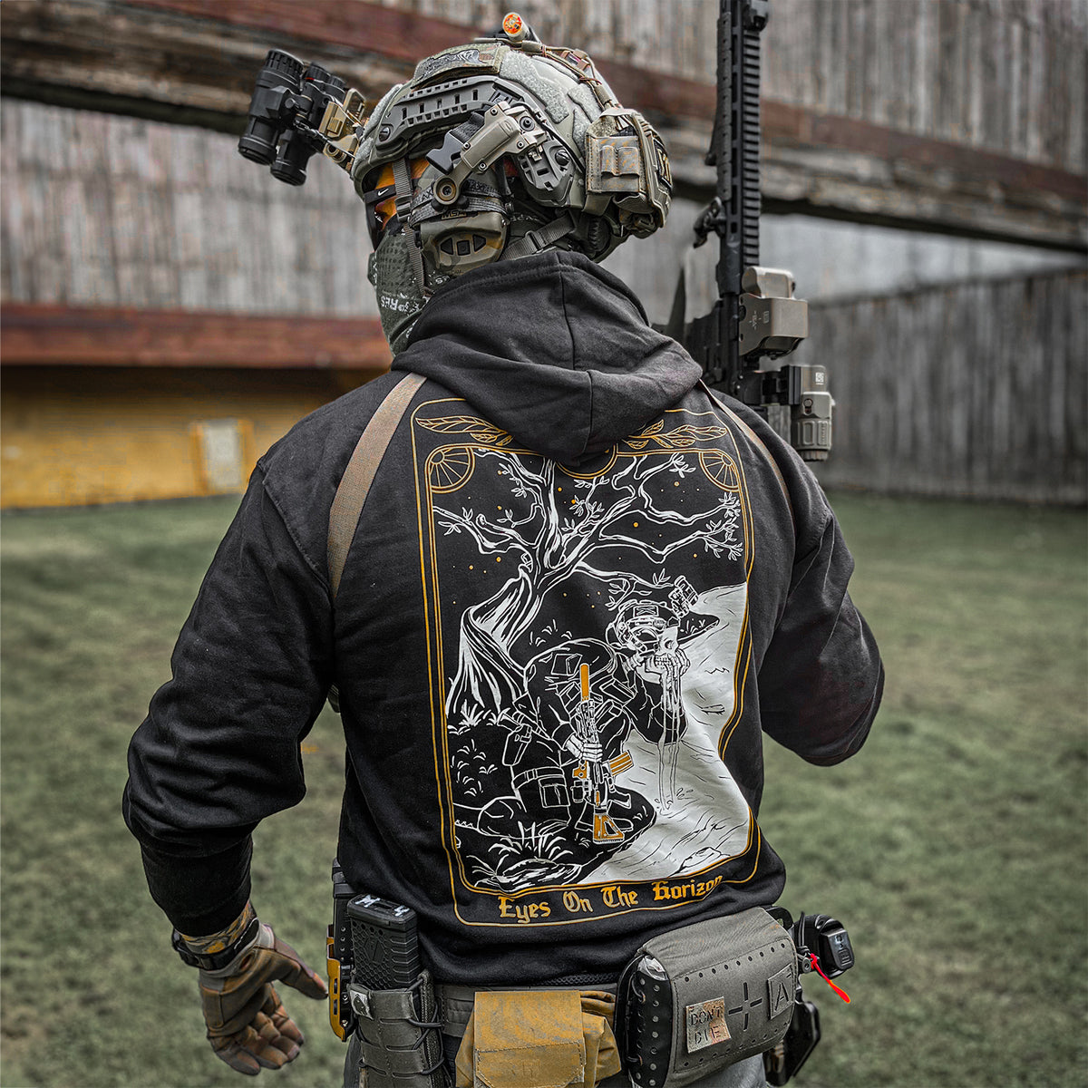 Person in taktischer Ausrüstung, die ein Gewehr hält, einen Kapuzenpullover mit illustriertem Design auf der Rückseite trägt und im Freien steht.
