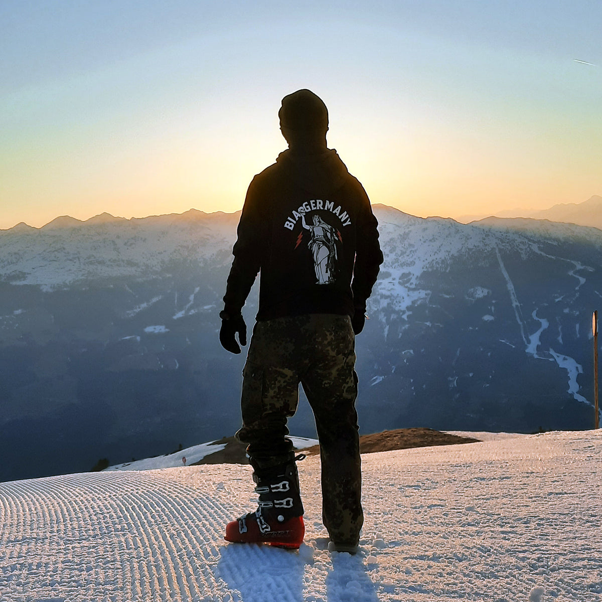Man from behind in the black Germania Hoodie on a snowy slope at sunrise