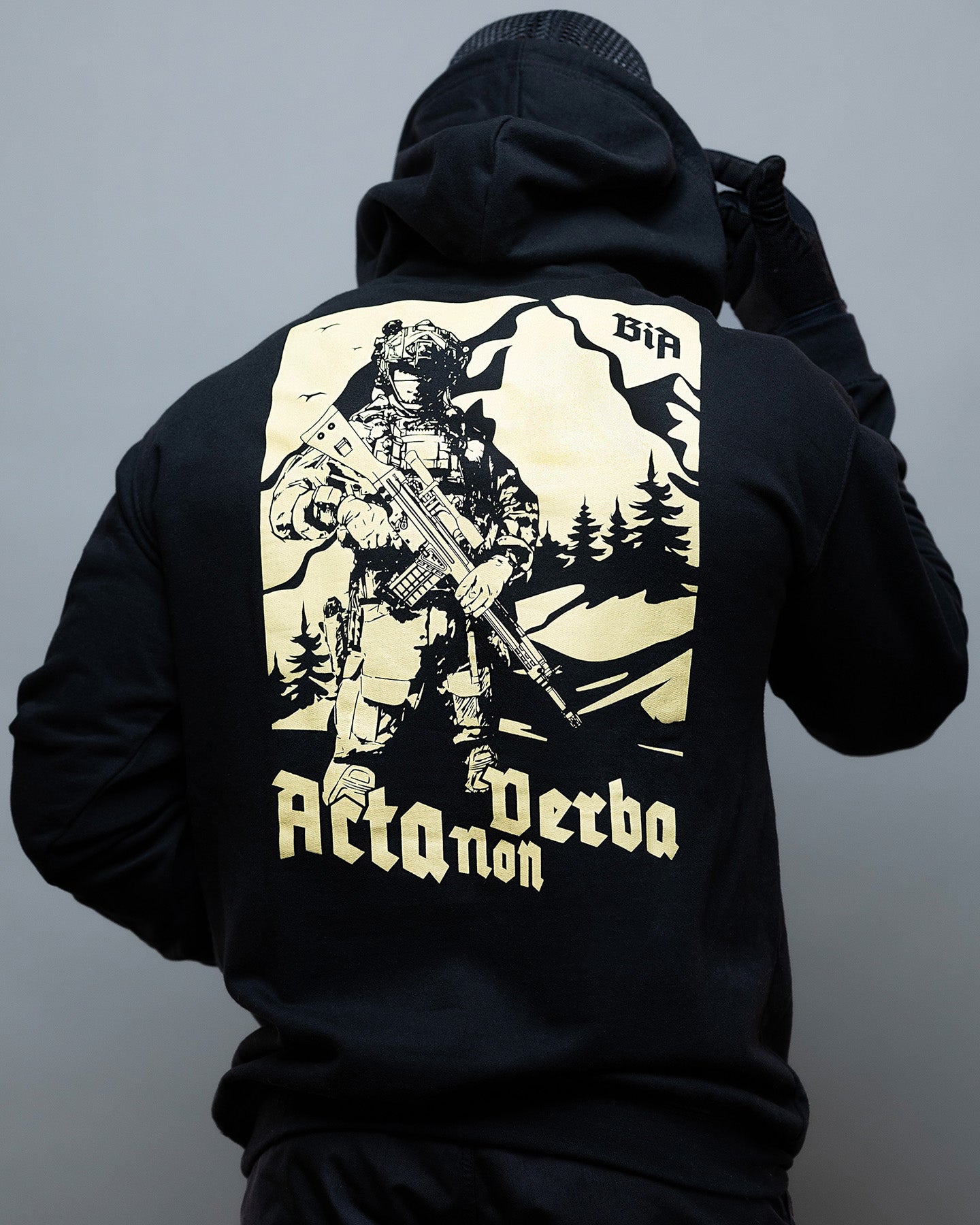 Person wearing a black hoodie with a graphic design on the back against a gray background - Acta Non Verba Hoodie