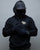 Person wearing a black hoodie with text, black cap, and gloves on a gray background - Acta Non Verba Hoodie