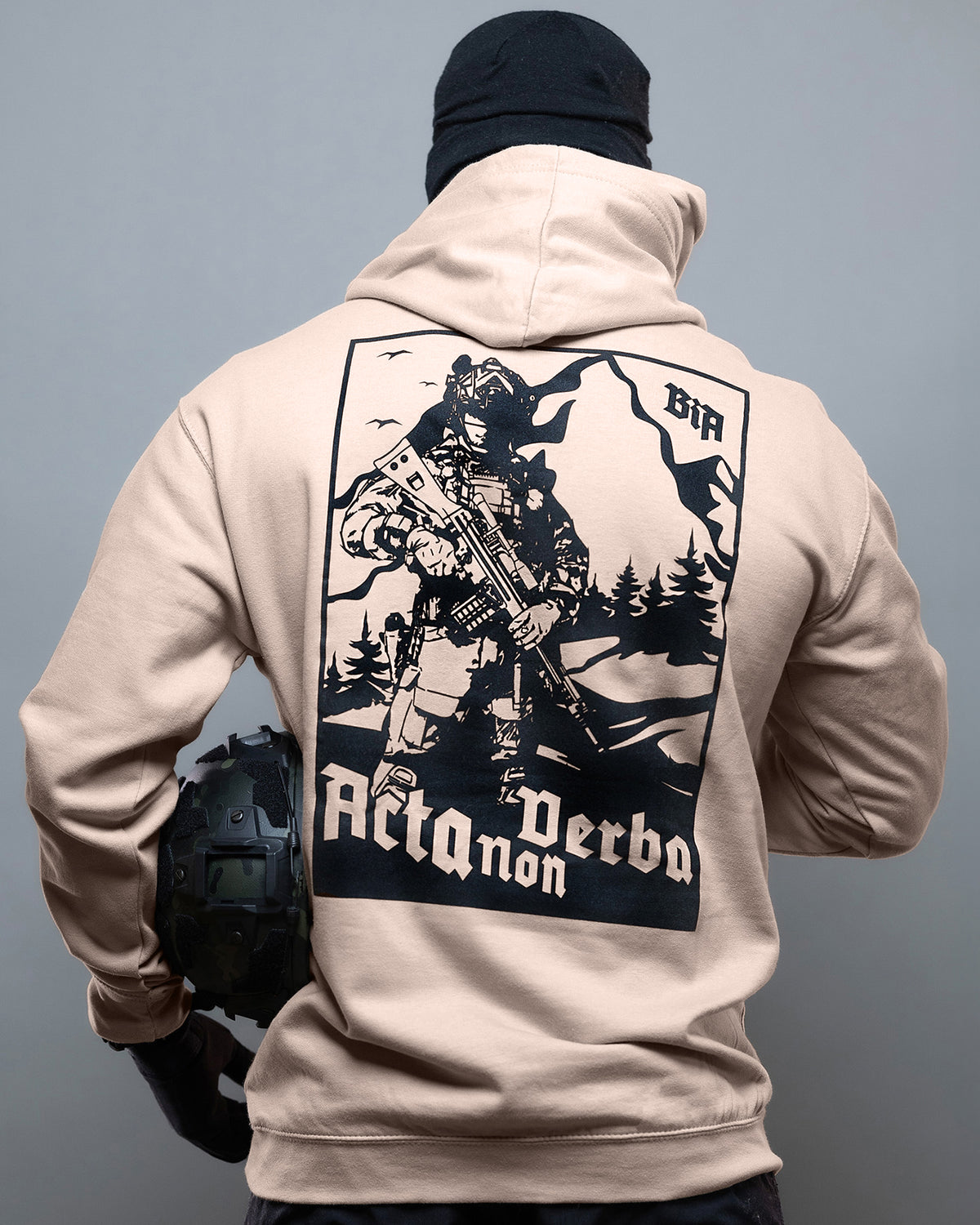 Person wearing a beige hoodie with a graphic design on the back against a gray background - Acta Non Verba Hoodie