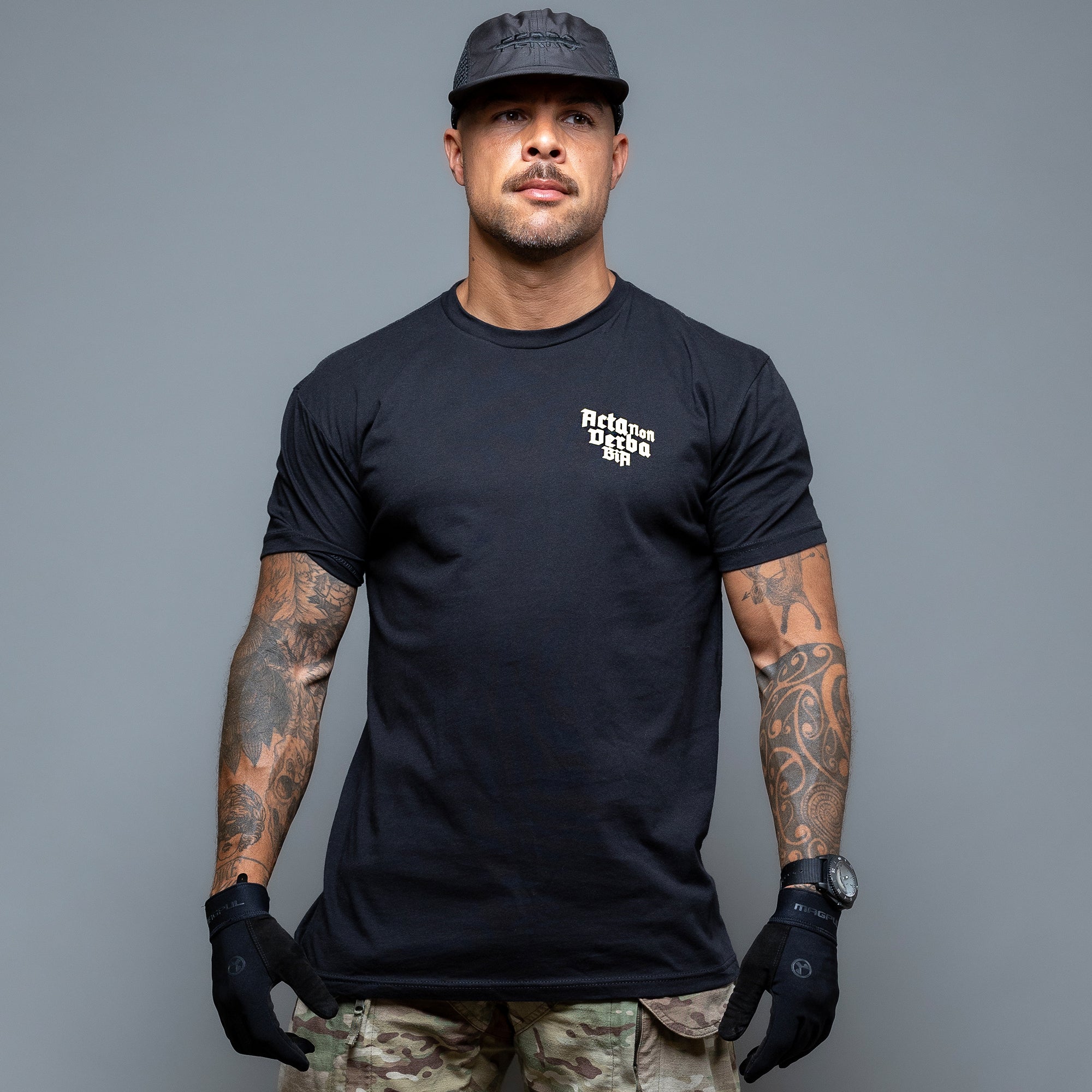 Ein Mann mit schwarzer Kappe und sichtbaren Tattoos unter seinem Acta Non Verba T-Shirt von Brothers in Arms steht selbstbewusst vor einem grauen Hintergrund. Das Funktionsshirt und die Handschuhe betonen „Acta Non Verba“, das in jede Faser seiner atmungsaktiven Kleidung eingraviert ist.