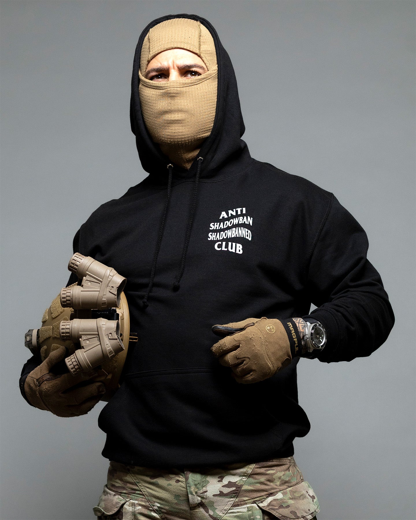 Person wearing a black hoodie with text, beige gloves, and a balaclava on a gray background. #color_black