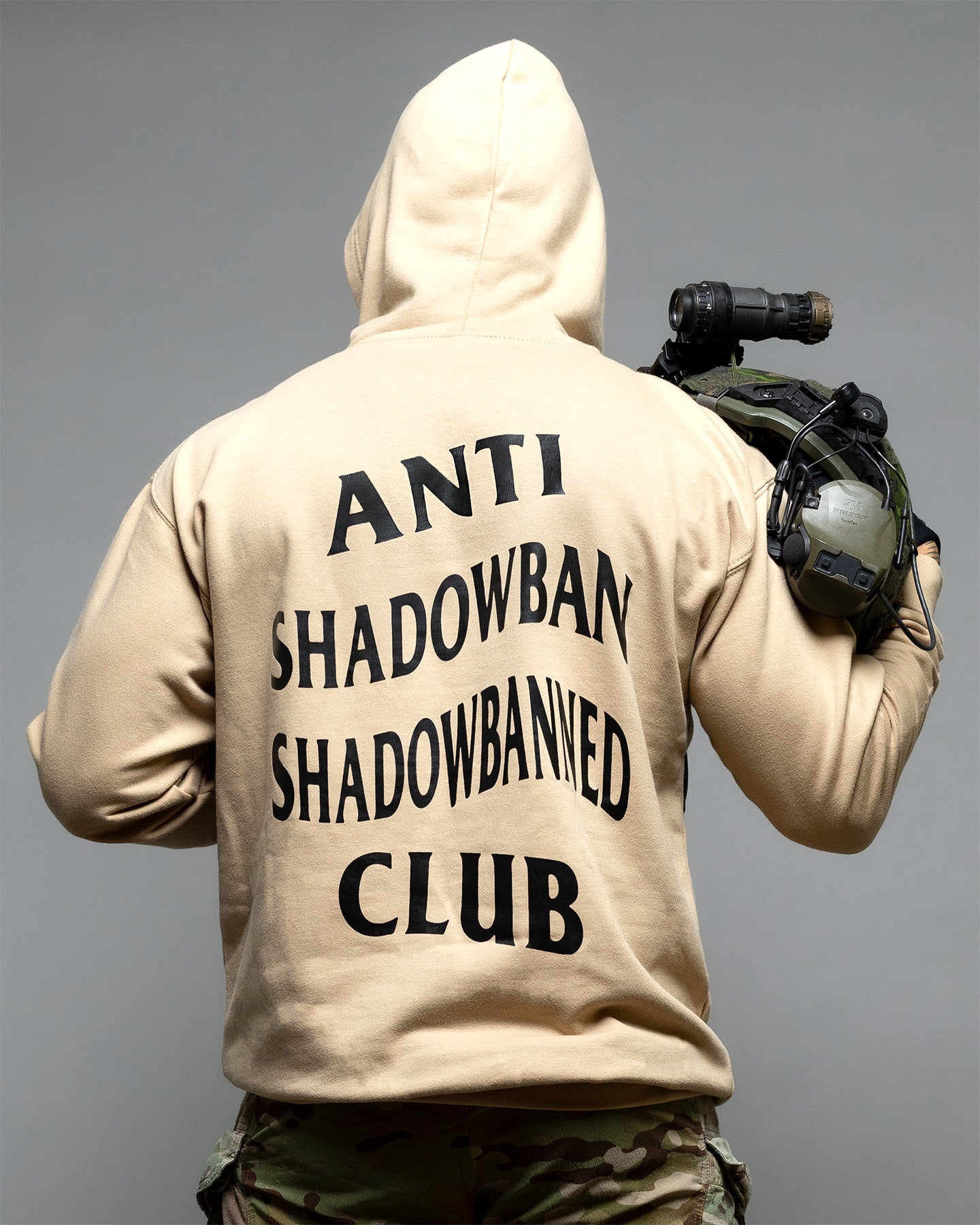 Person wearing a beige hoodie with 'ANTI SHADOWBAN SHADOWBANNED CLUB' text on the back, holding a weapon. #color_sand