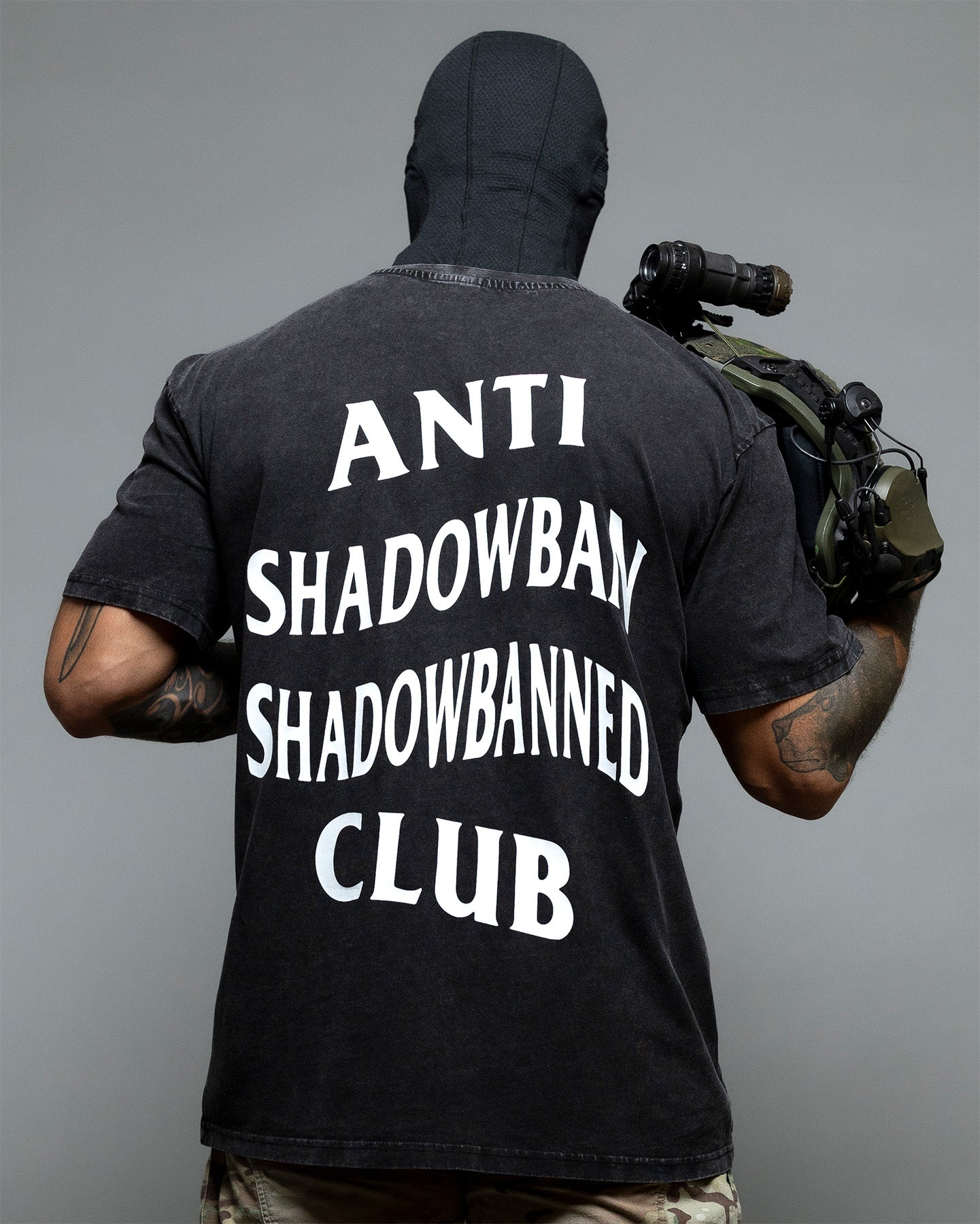 Person wearing a black t-shirt with 'ANTI SHADOWBAN SHADOWBANNED CLUB' text on the back, holding a helmet. #color_black
