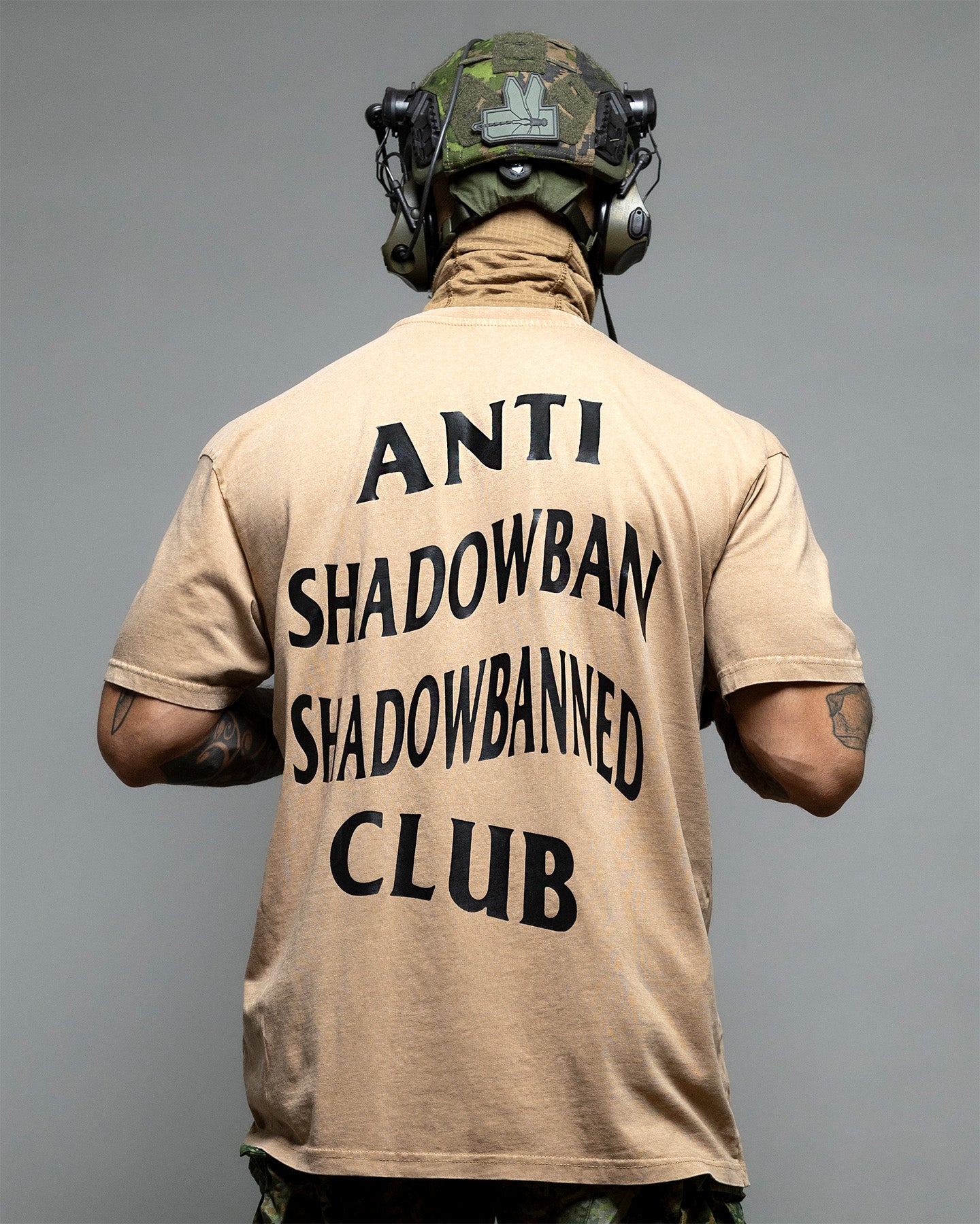 Person wearing a beige shirt with 'ANTI SHADOWBAN SHADOWBANNED CLUB' text on the back, against a gray background. #color_coyote
