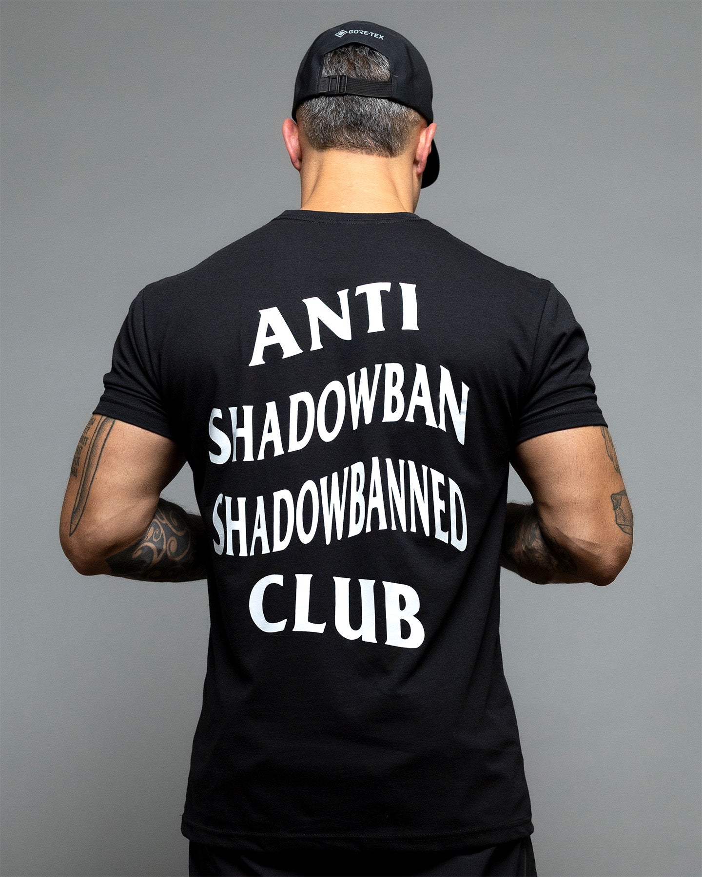 Person wearing a black t-shirt with 'ANTI SHADOWBAN SHADOWBANNED CLUB' text on the back against a gray background. #color_black