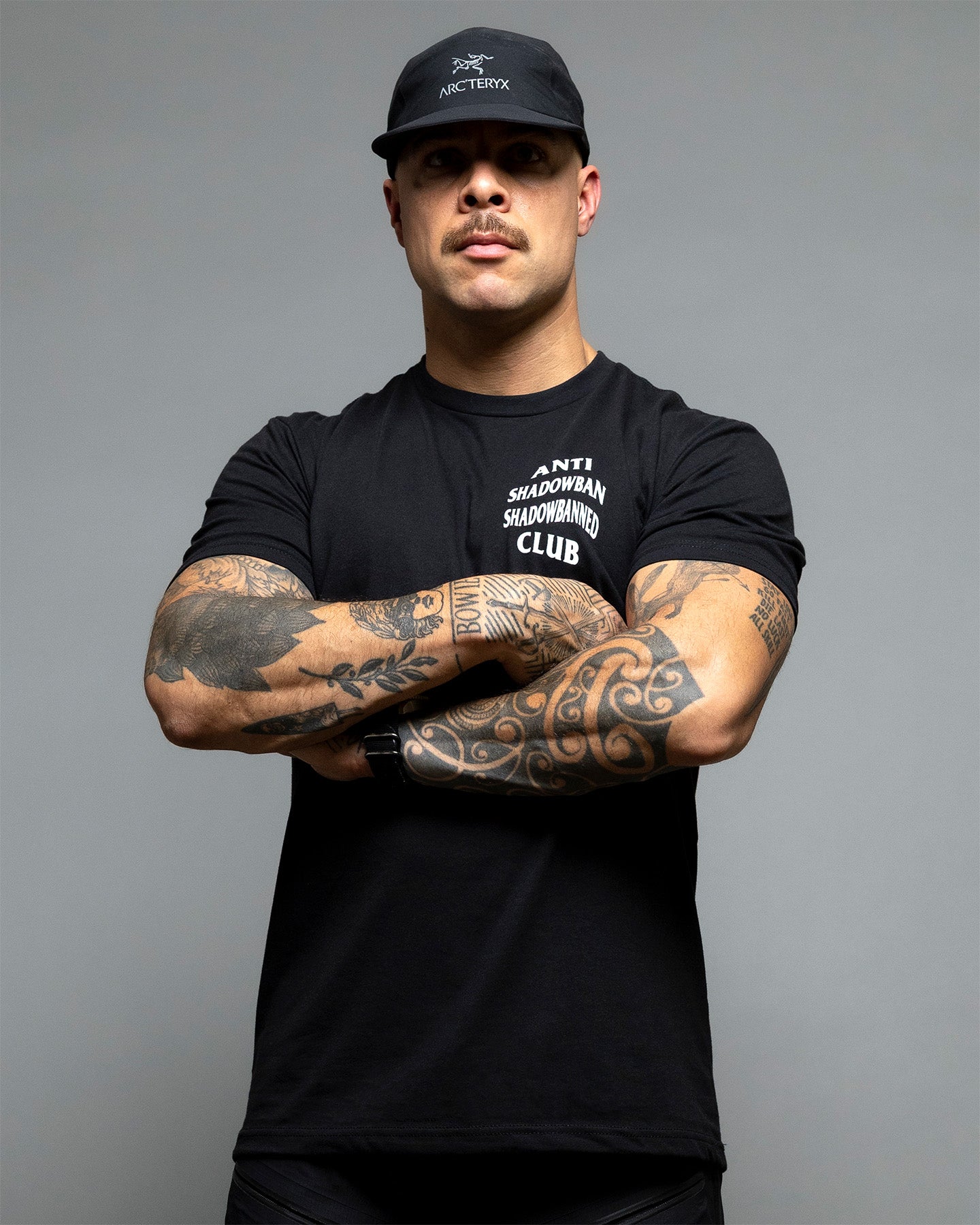 Man wearing a black t-shirt and cap with visible tattoos, standing against a gray background. Person wearing a black t-shirt with 'ANTI SHADOWBAN SHADOWBANNED CLUB' text on the back against a gray background. #color_black
