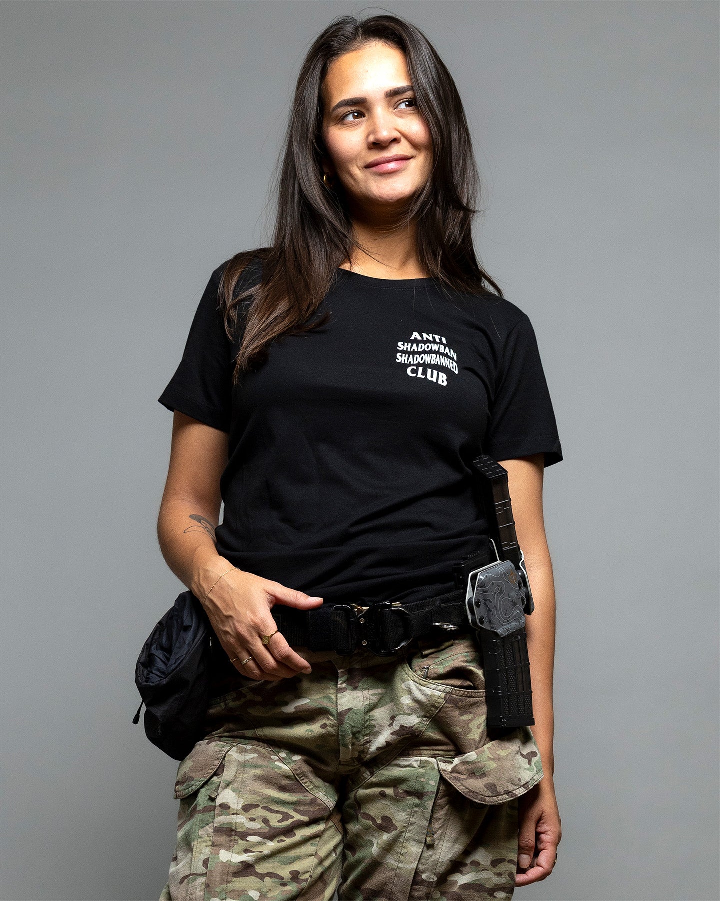 Person wearing a black t-shirt with text, camouflage pants, and carrying a bag on a gray background. #color_black