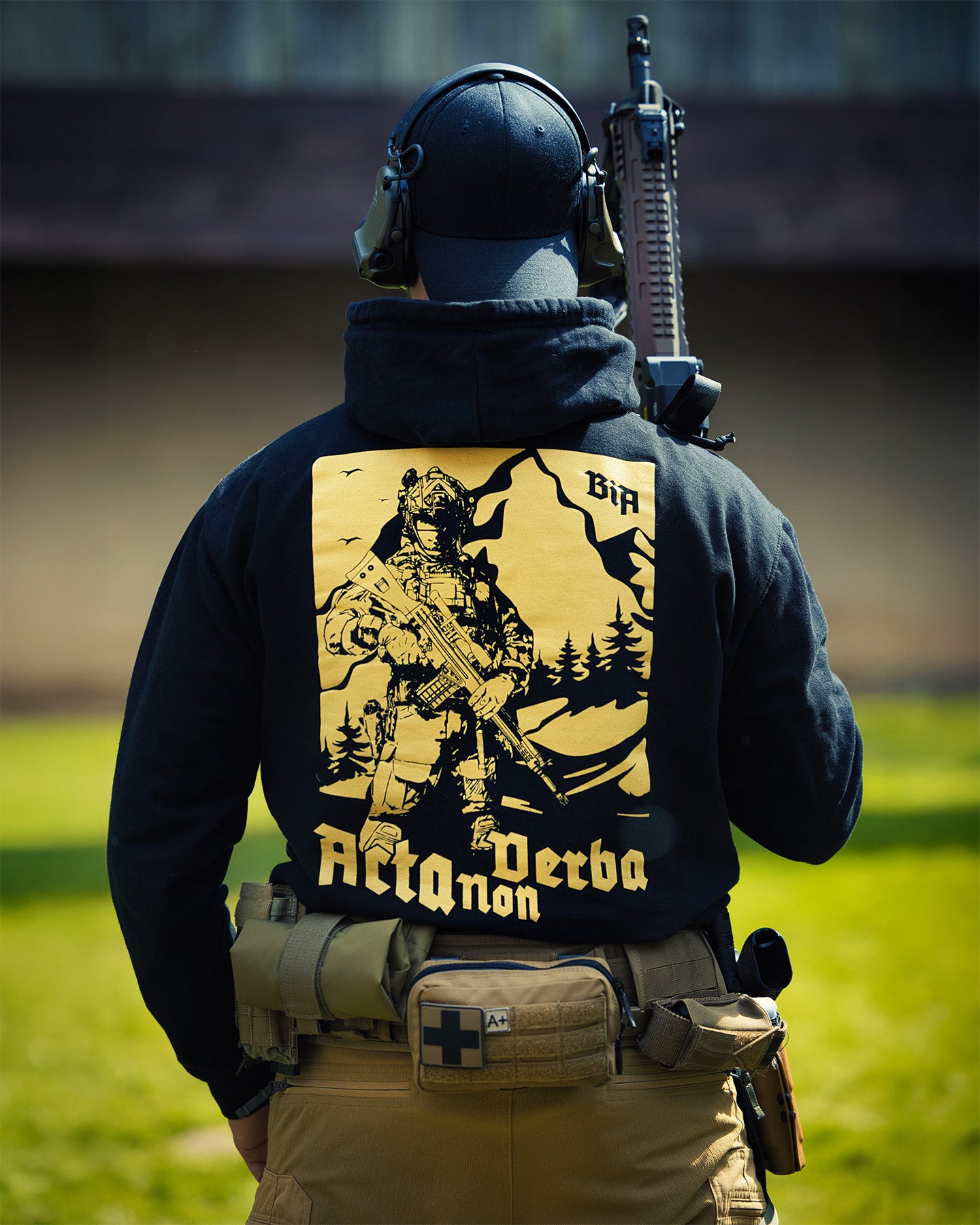 Person wearing a tactical outfit with a visible brand logo on the back, standing in an outdoor setting - Acta Non Verba Hoodie