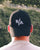 Person wearing a black cap with a logo, facing away from the camera with a blurred green background.