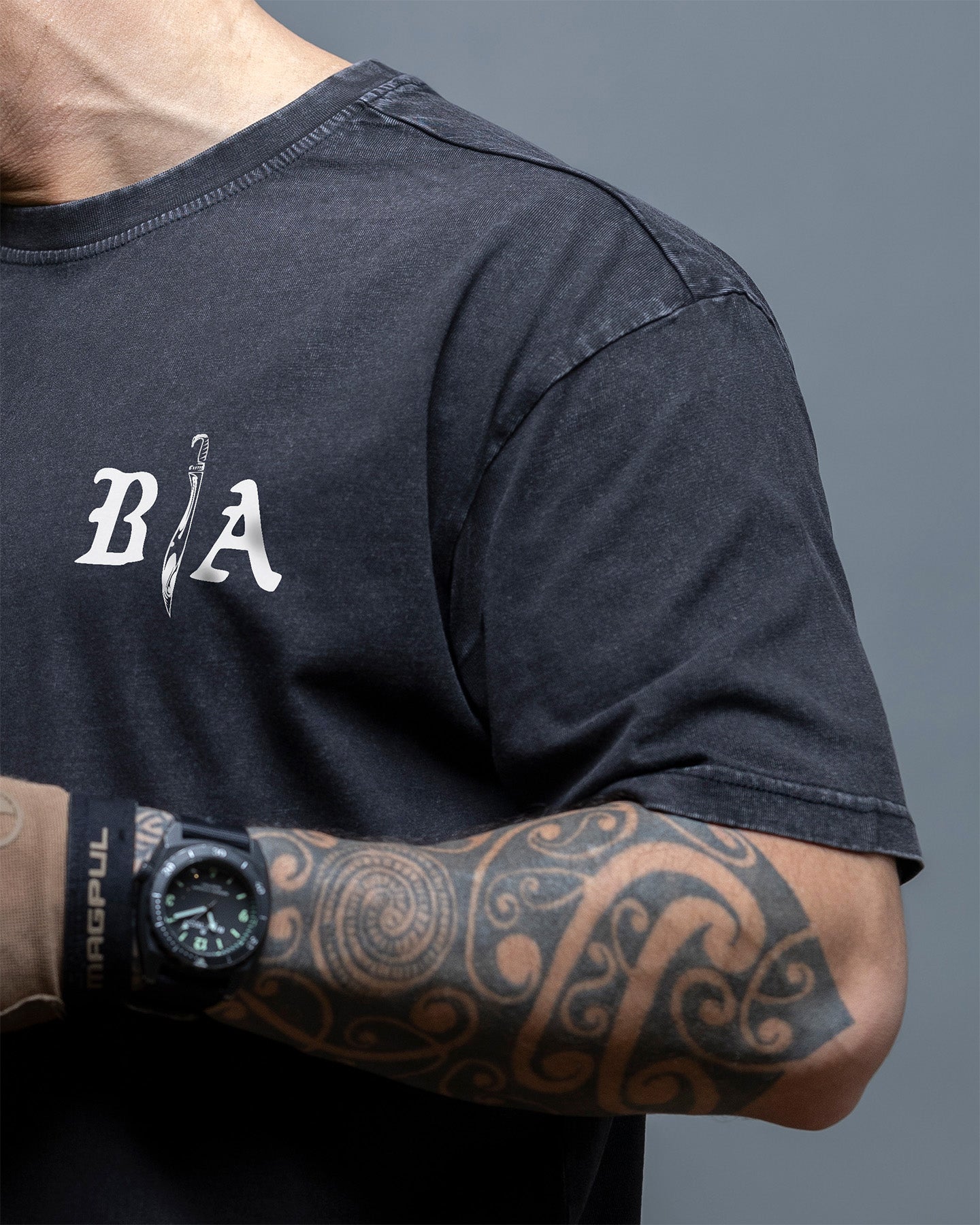Person wearing a black t-shirt with a logo and a tattooed arm, on a gray background. #color_black
