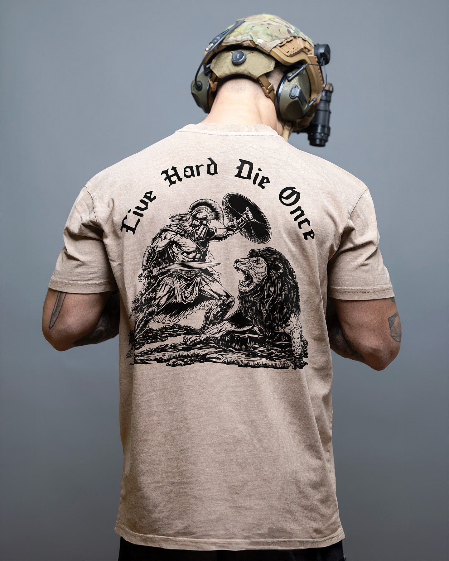 Person wearing a beige t-shirt with a graphic design and text on the back, against a gray background. #color_coyote
