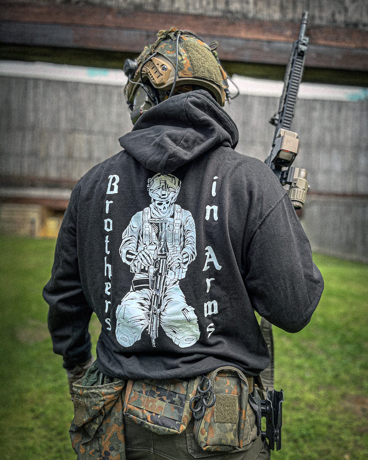 Man from behind in the black Brothers in Arms Hoodie with plate carrier and rifle in the field