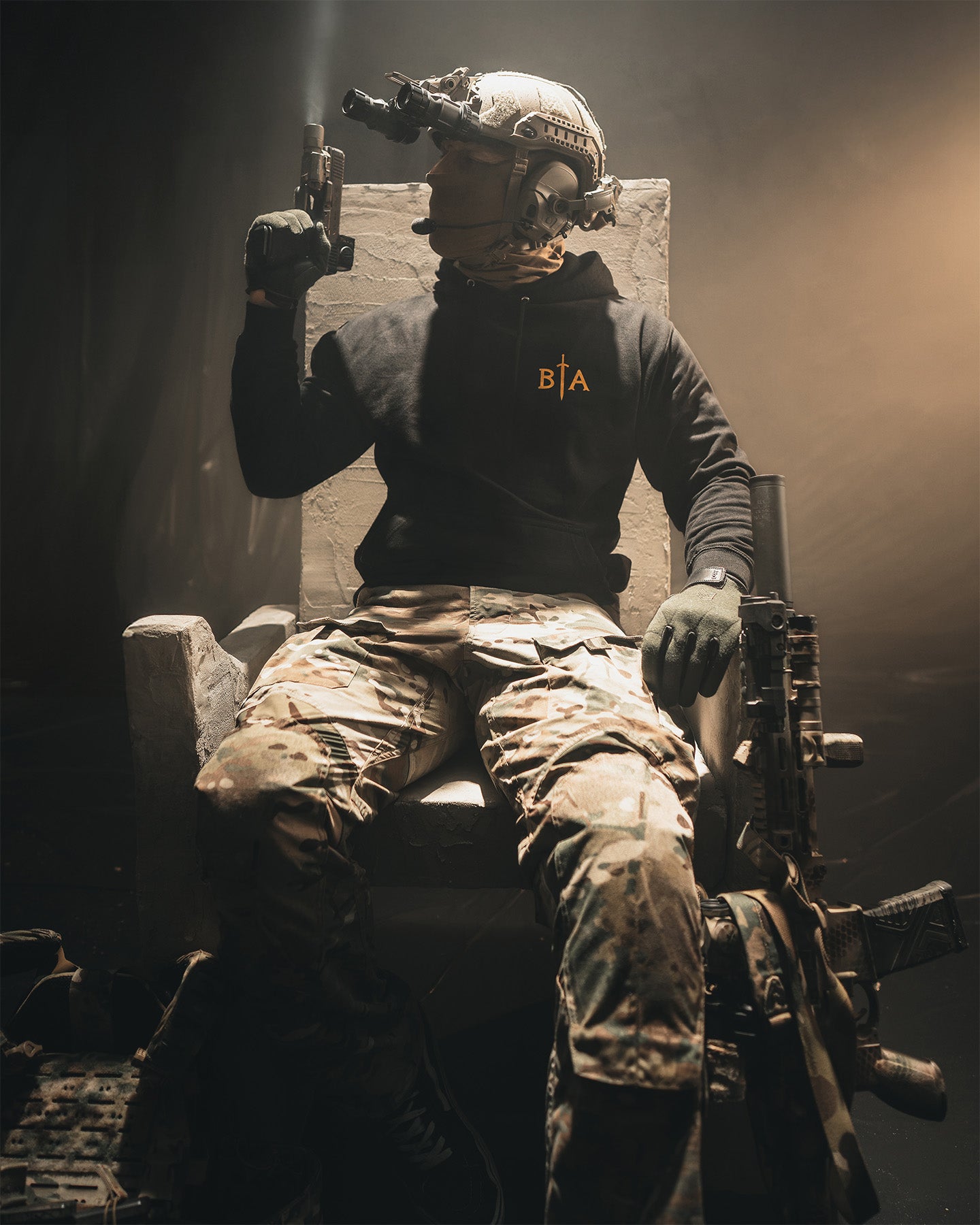 Person in tactical gear sitting on a chair with weapons, wearing a hoodie with a logo. #color_black #in_field