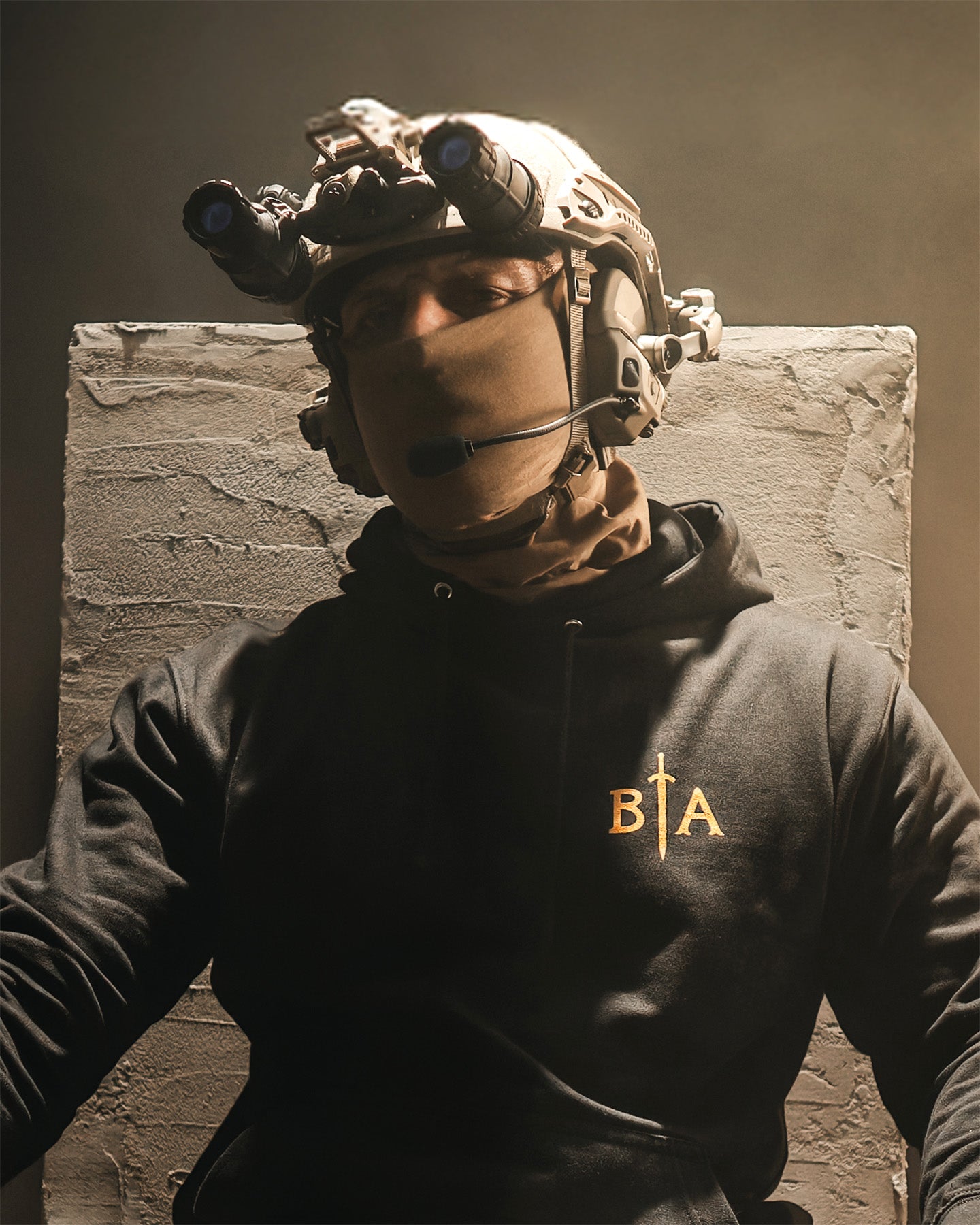 Person wearing tactical gear and a hoodie with a logo, standing against a textured wall. #color_black #in_field
