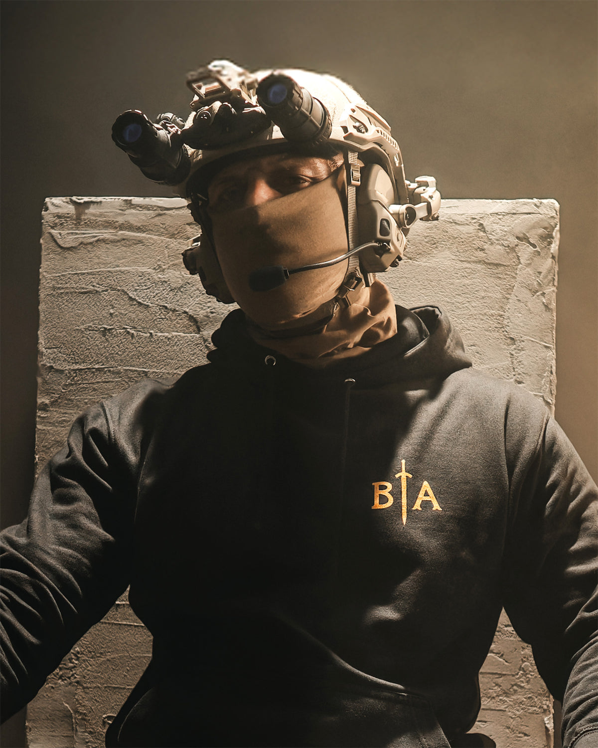 Person in a helmet with night vision seated against a stone block, wearing the black No Fear Hoodie