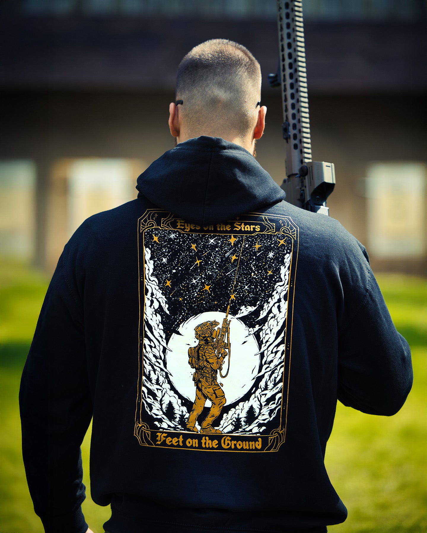 Man from behind in the black Eyes On The Stars Hoodie with a rifle, tarot card back print #color_black #in_field