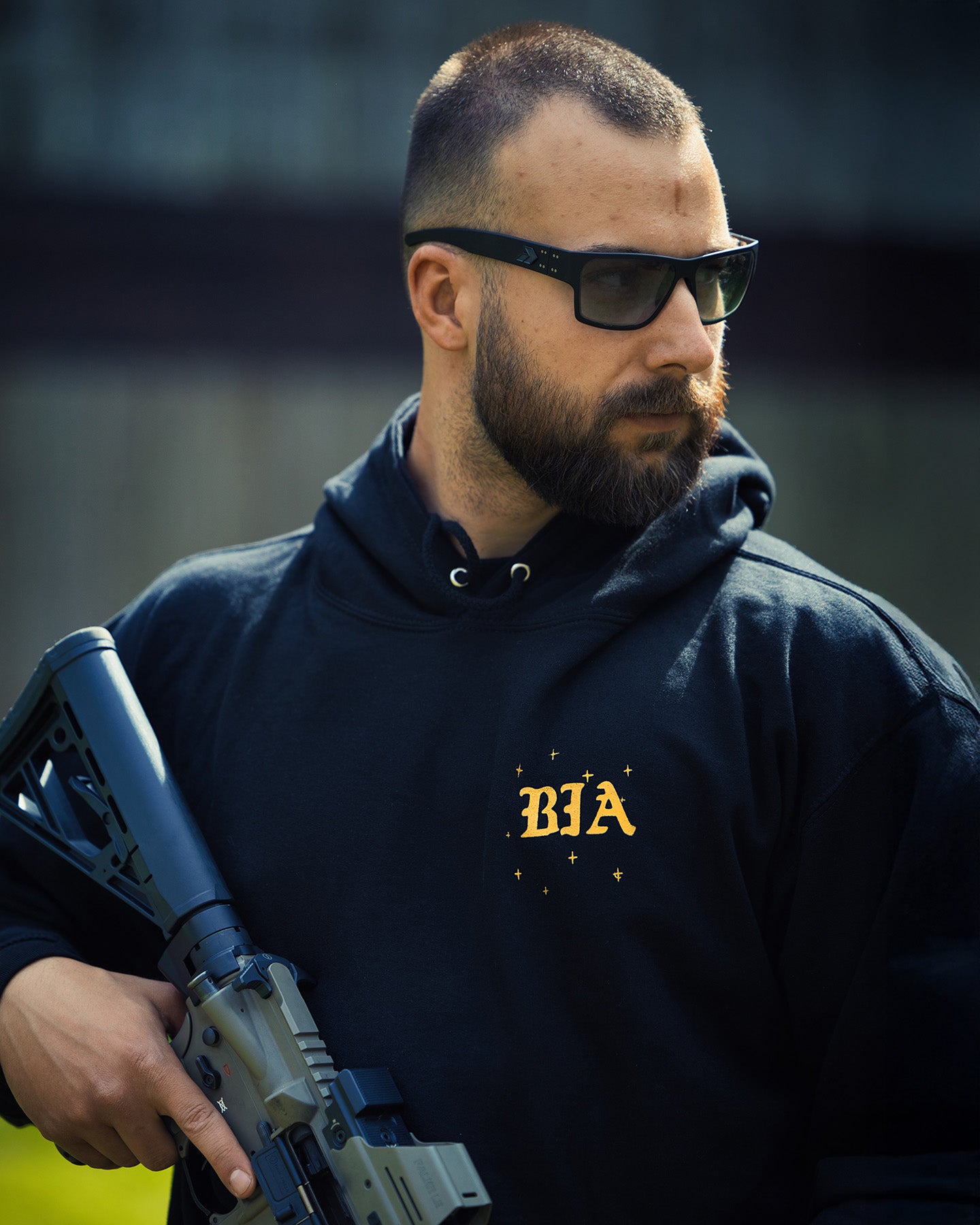Man in sunglasses holding a rifle, wearing the black Eyes On The Stars Hoodie #color_black #in_field