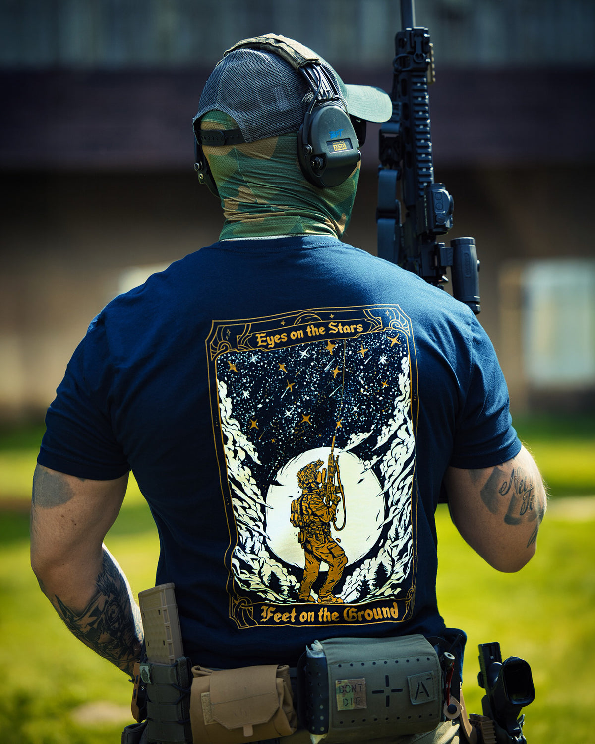 Man from behind in the navy Eyes On The Stars T-Shirt with a rifle on a training ground