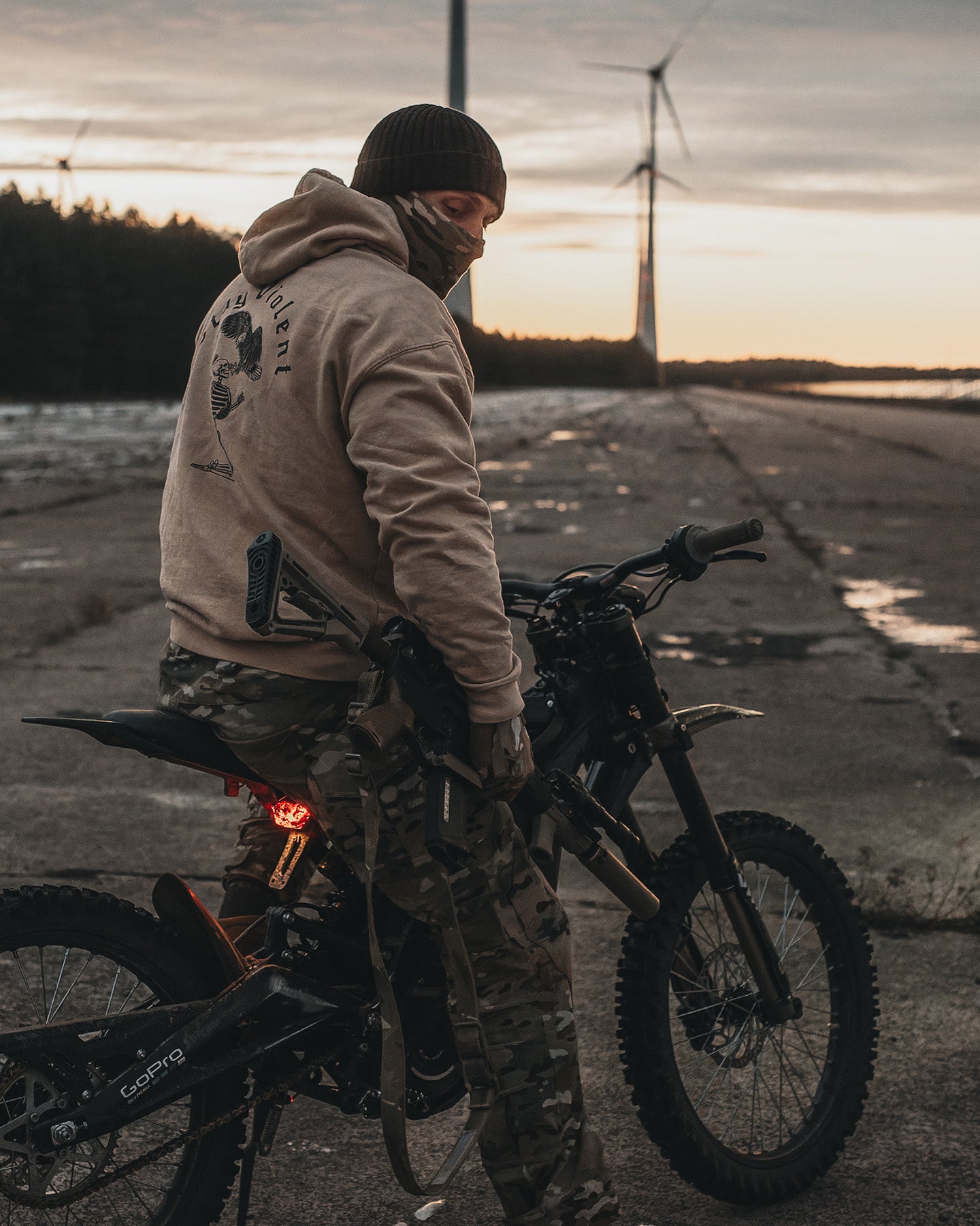 Man on a motorcycle in the sand Stay Violent Hoodie at dusk in front of wind turbines #color_sand #in_field