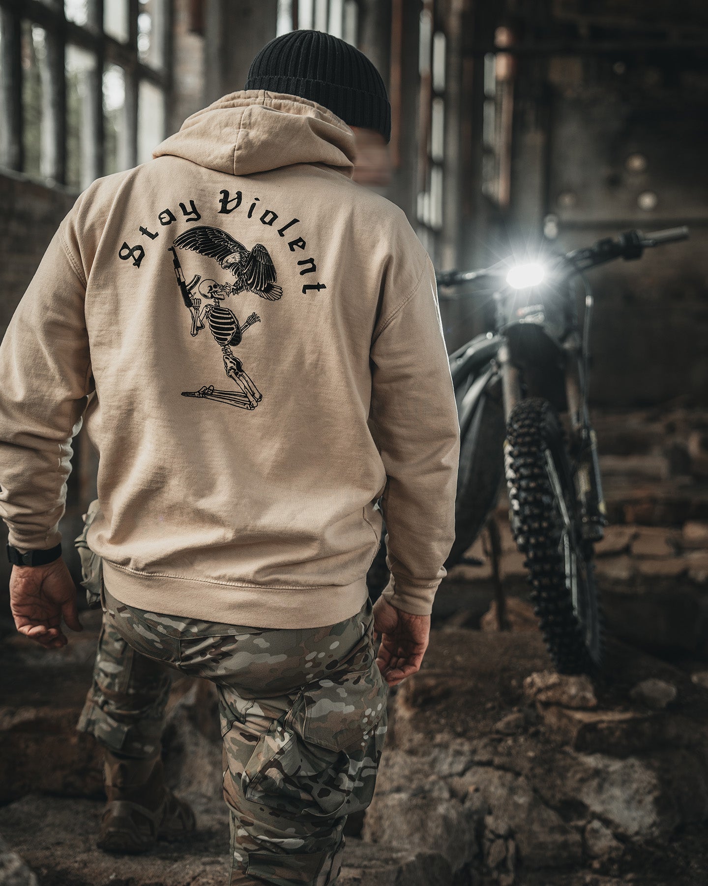 Man from behind in the sand Stay Violent Hoodie next to a motorcycle in an abandoned building #color_sand #in_field