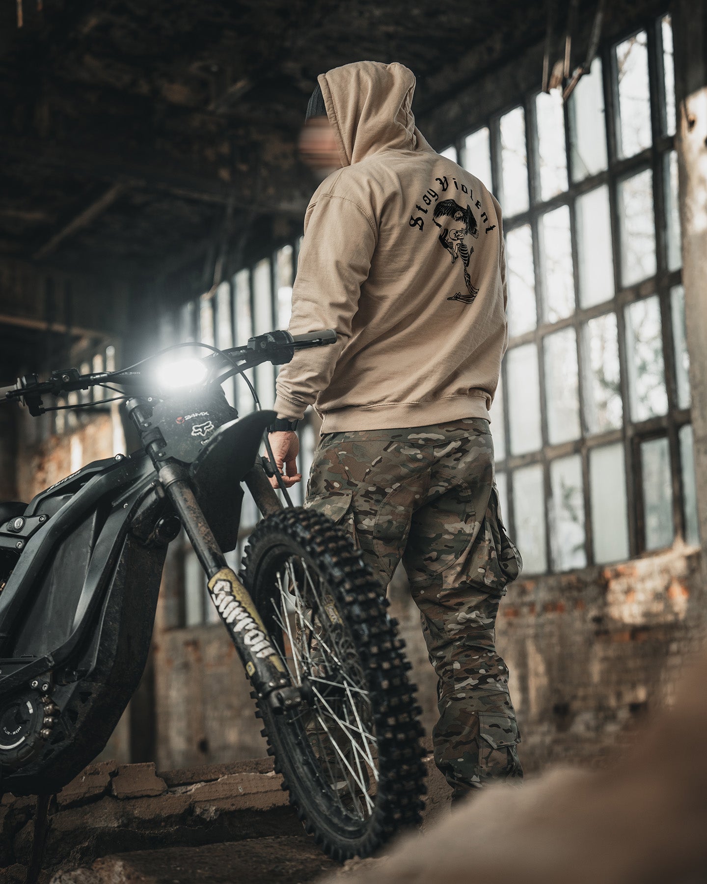 Man in the sand Stay Violent Hoodie beside a motorcycle in an abandoned building #color_sand #in_field