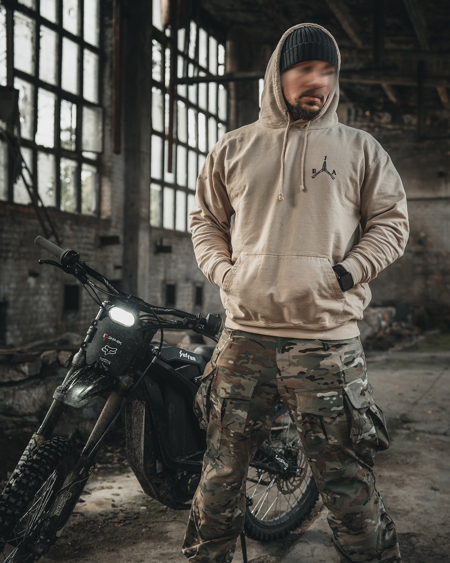 Man in a beanie and the sand Stay Violent Hoodie standing next to a motorcycle #color_sand #in_field