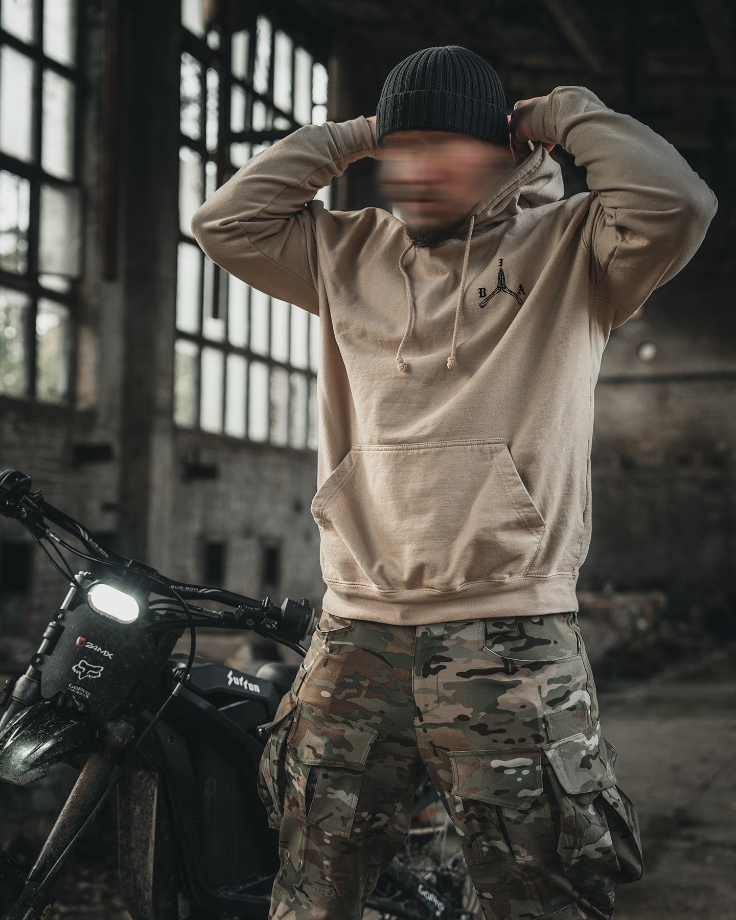 Man in the sand Stay Violent Hoodie with his hands behind his head beside a motorcycle #color_sand #in_field